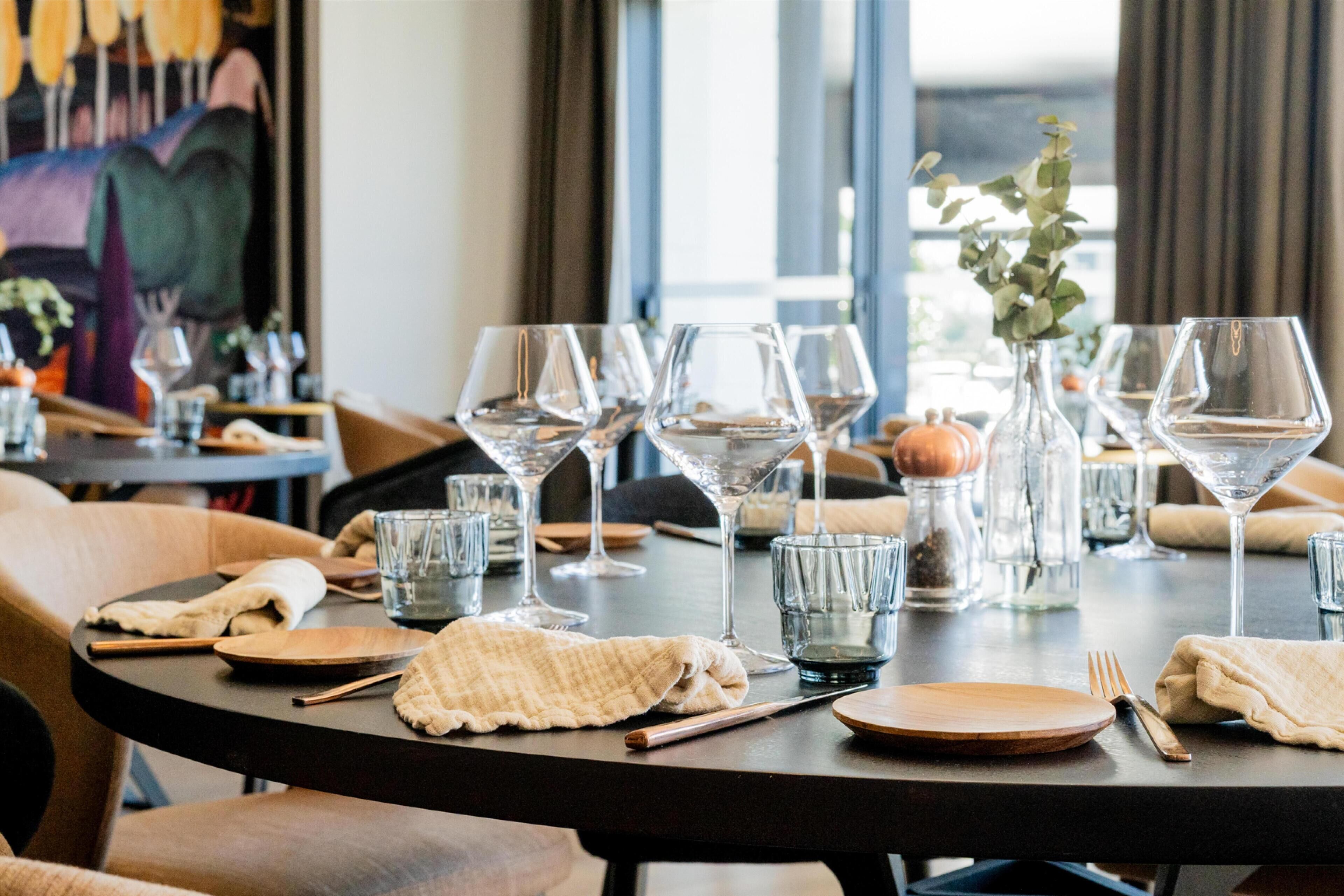 A dining table set with wine glasses, plates, and cutlery, with a view of the cityscape through large windows.