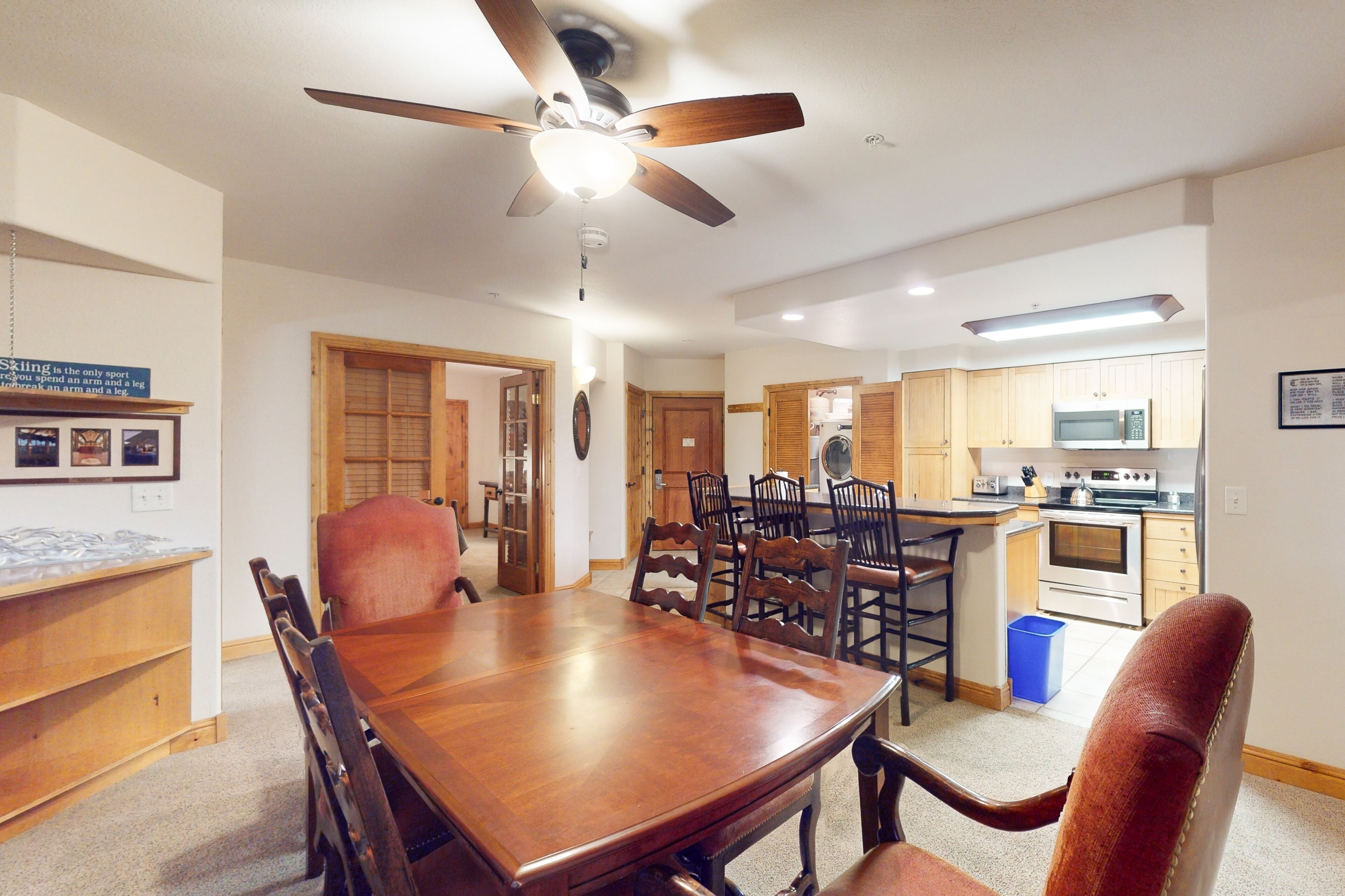 A dining area with a wooden table and chairs, a ceiling fan, and a kitchen in the background.