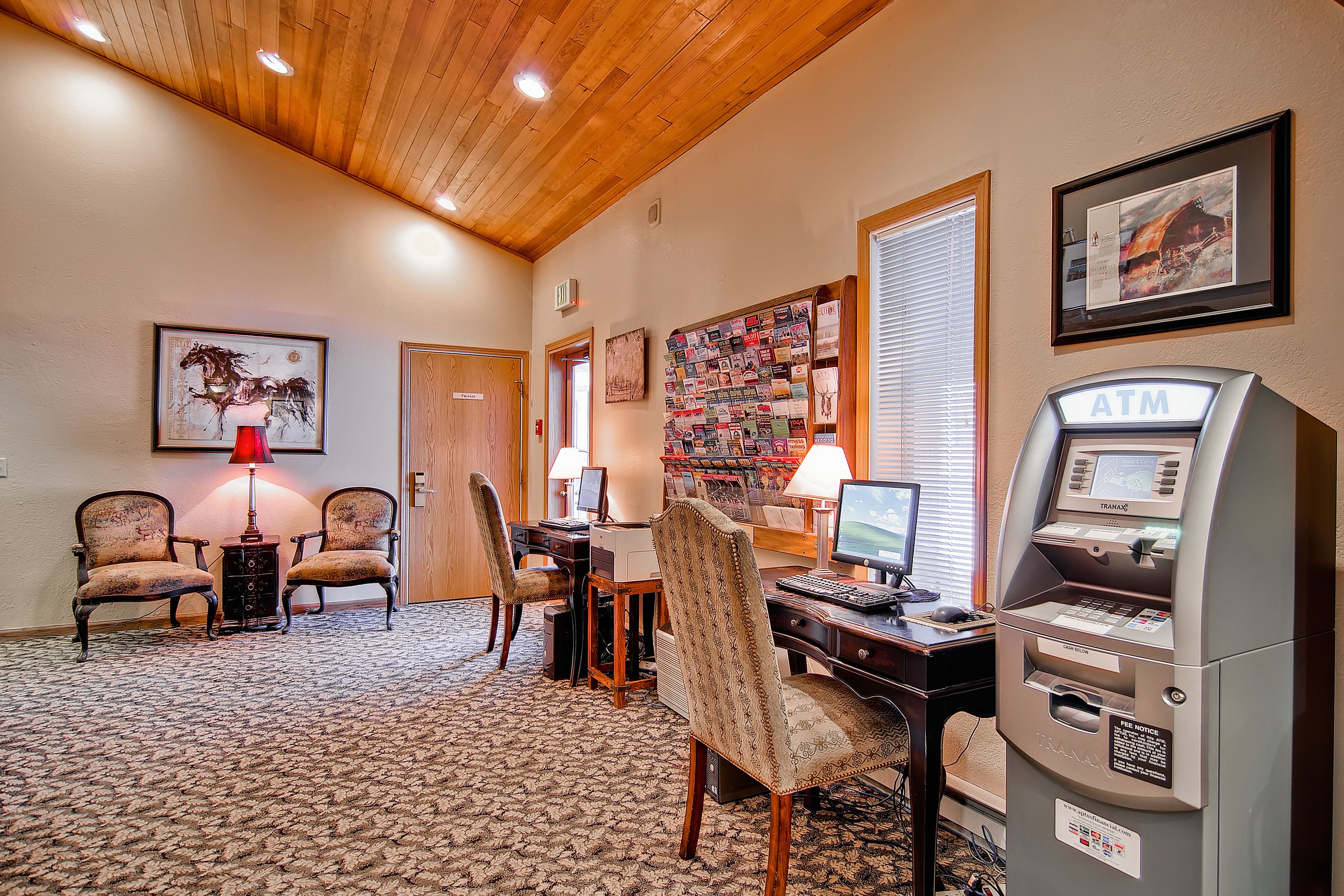 Une chambre d’hôtel avec un plafond en bois, un bureau avec un ordinateur, deux chaises, un distributeur automatique de billets et un tableau d’affichage.