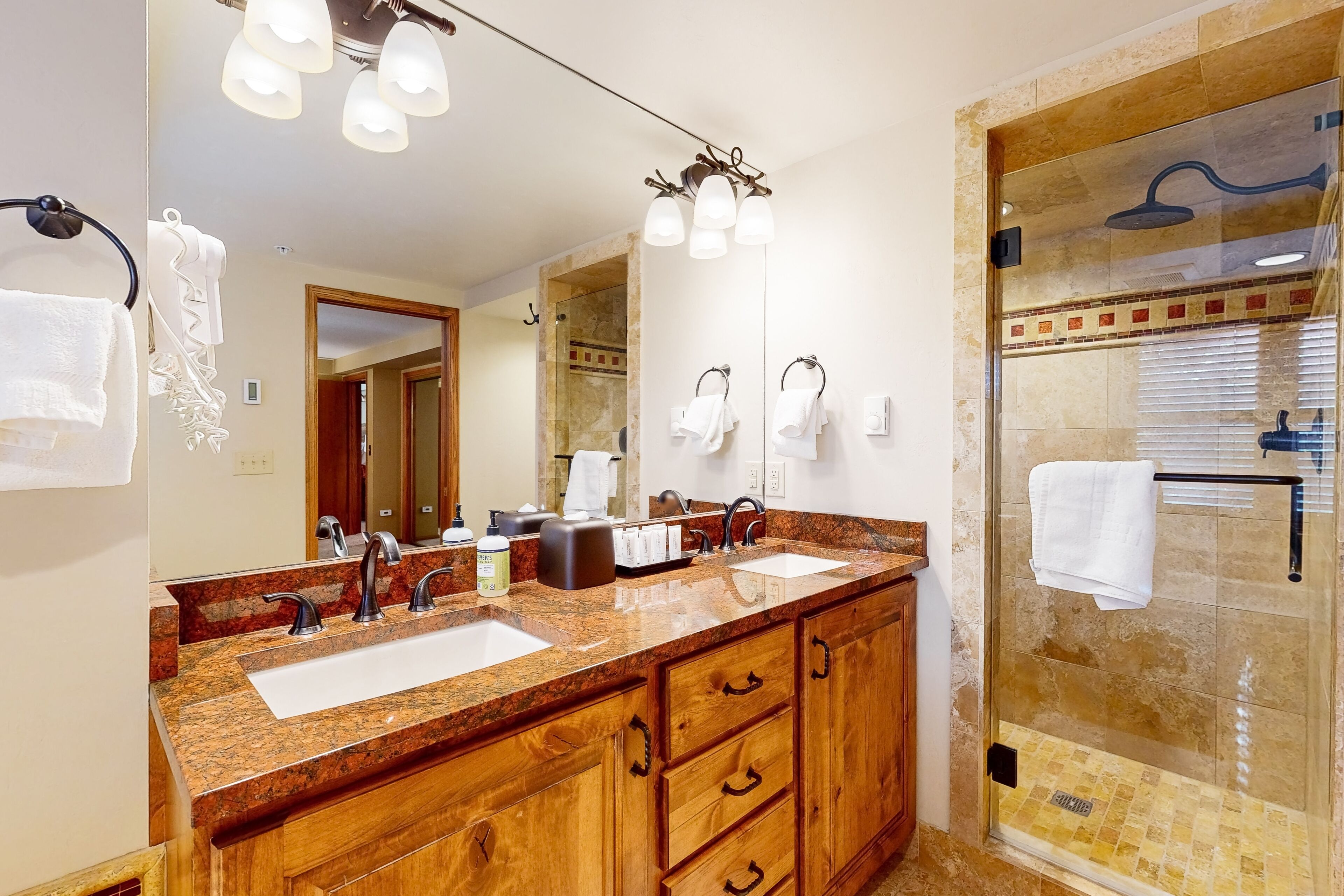 Un baño con encimera de granito, lavamanos dobles, un espejo grande y cabina de ducha con mampara de vidrio.