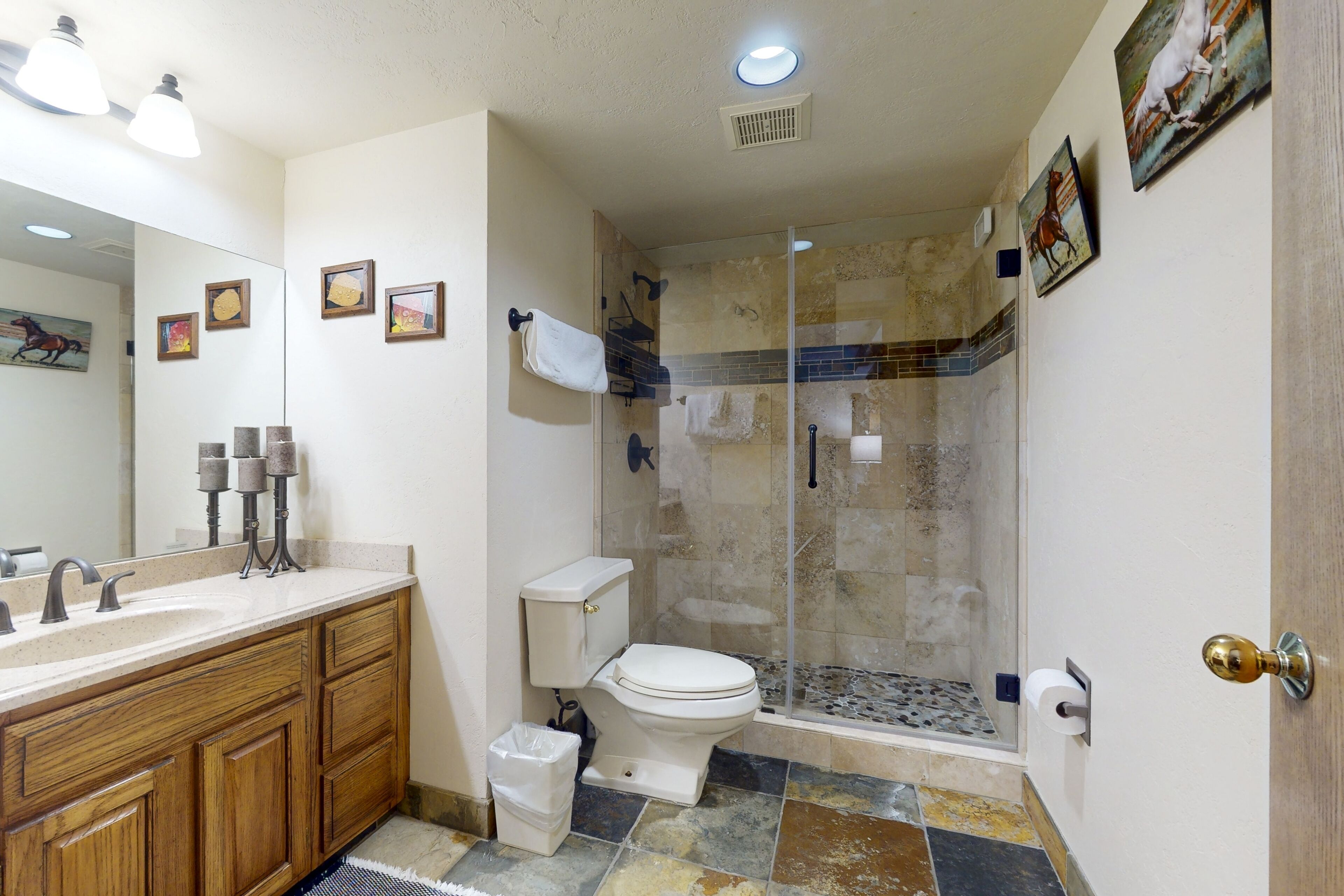 Un baño con tocador de madera, inodoro, ducha con puerta de vidrio y espejo con fotografías enmarcadas.