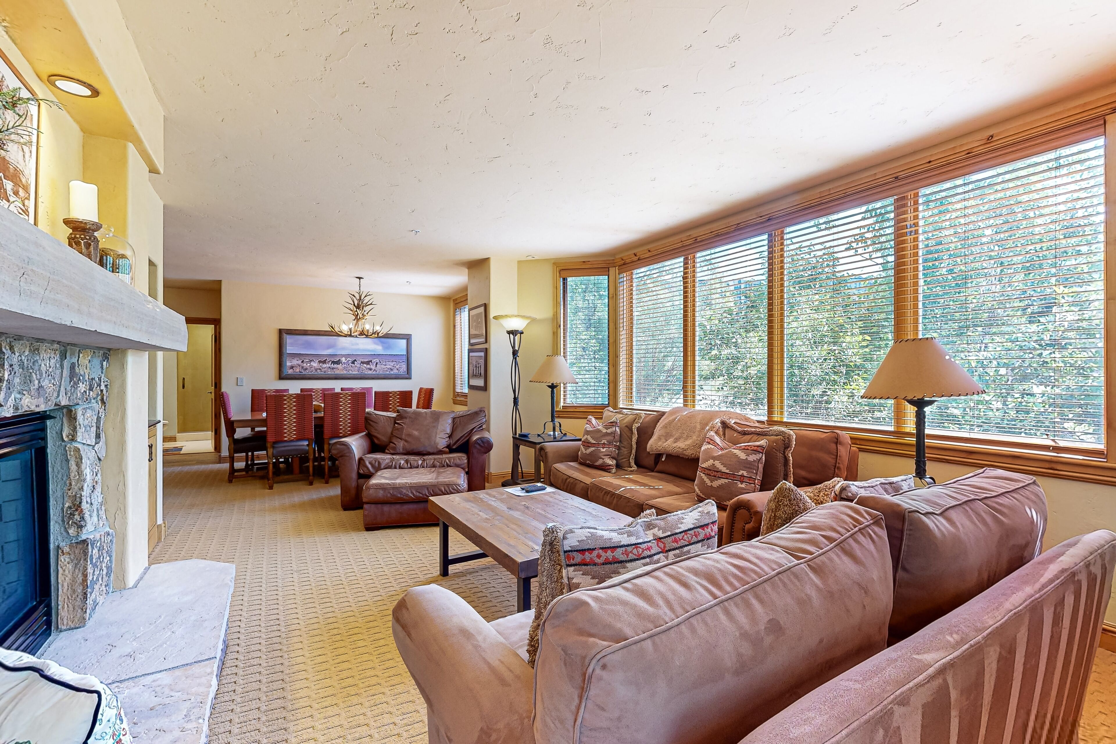 A spacious living room with a fireplace, leather sofas, a coffee table, and large windows with blinds.