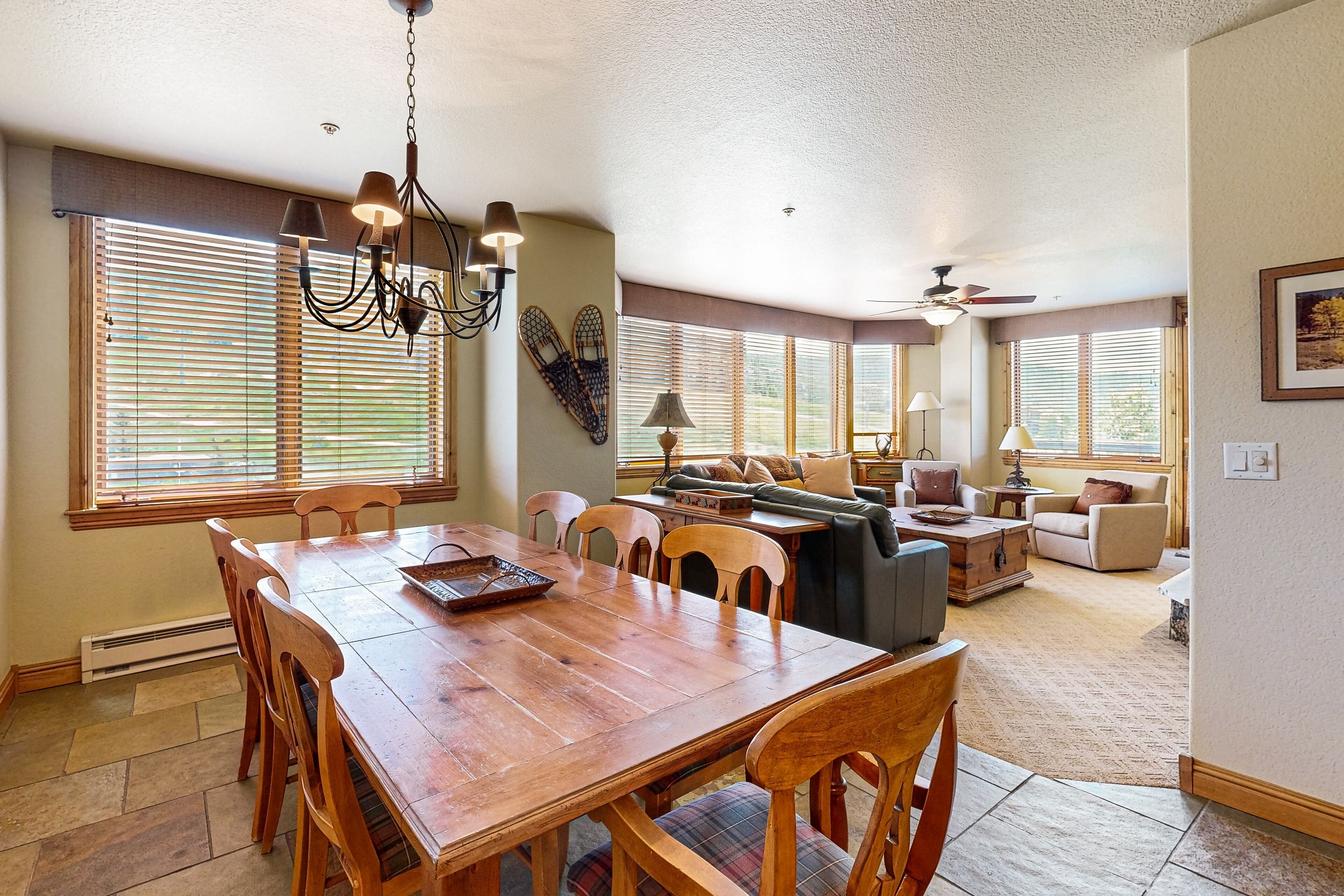 A spacious living area with a wooden dining table, comfortable seating, and large windows with blinds.