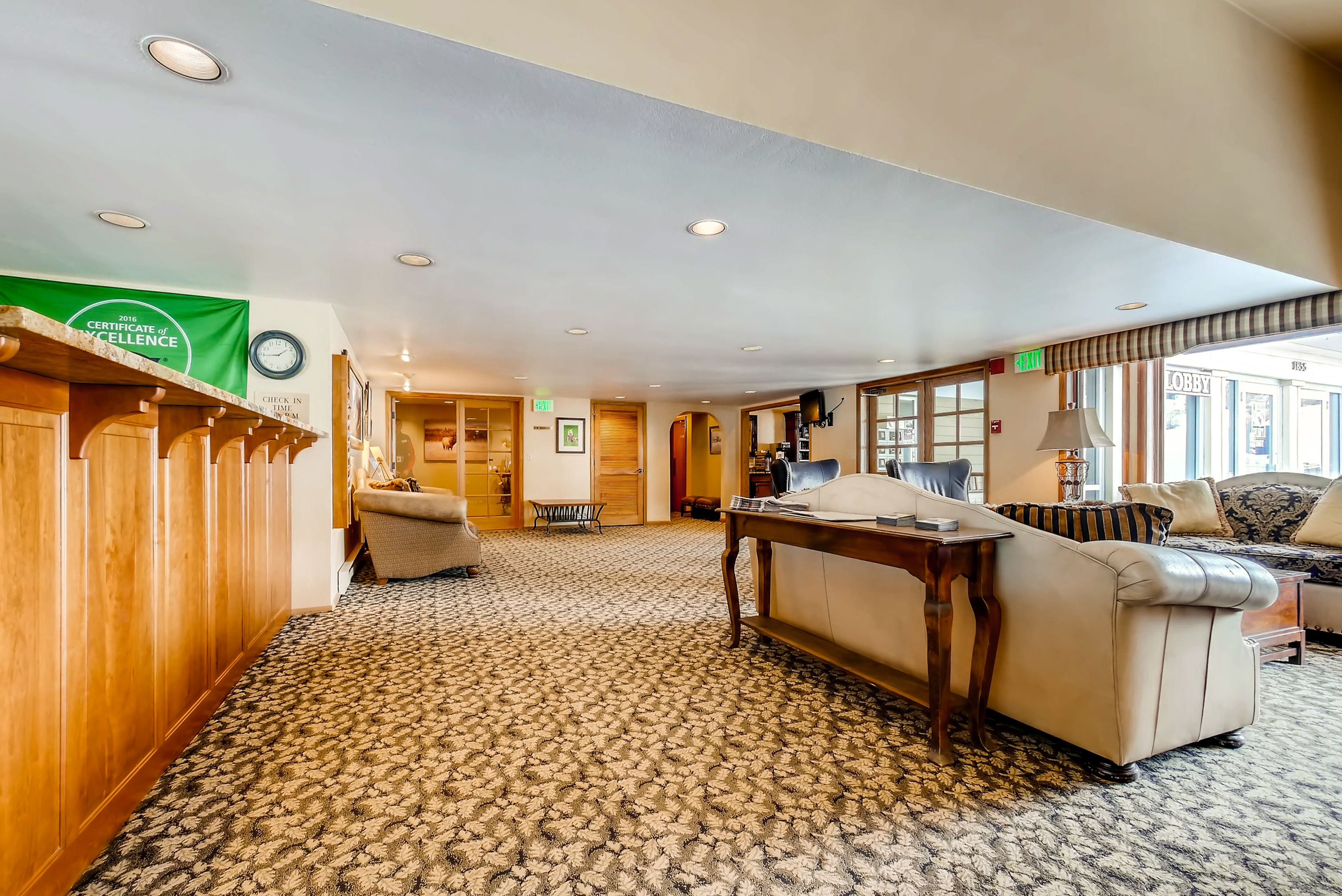 A hotel lobby with a patterned carpet, wooden reception desk, seating area with sofas, and a clock on the wall.