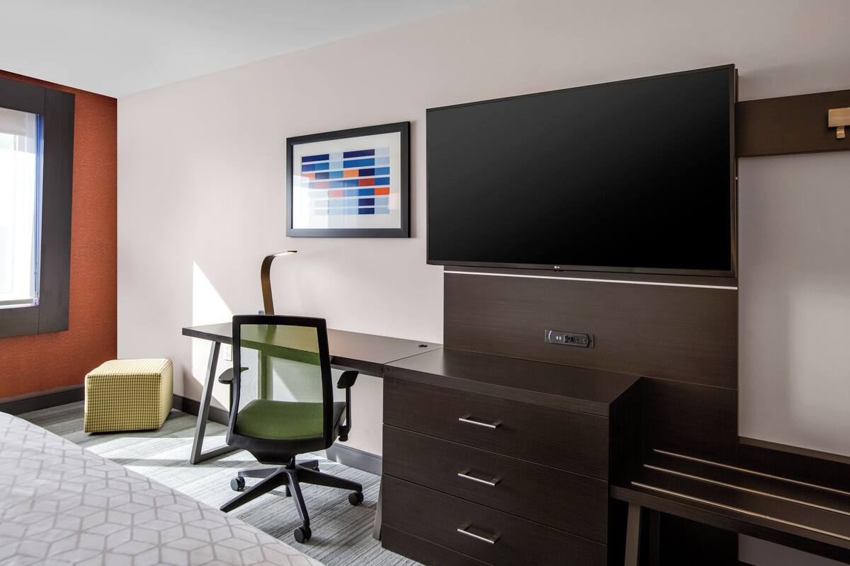 A modern hotel room with a large flat-screen TV, a desk with drawers, a green office chair, and a small ottoman.