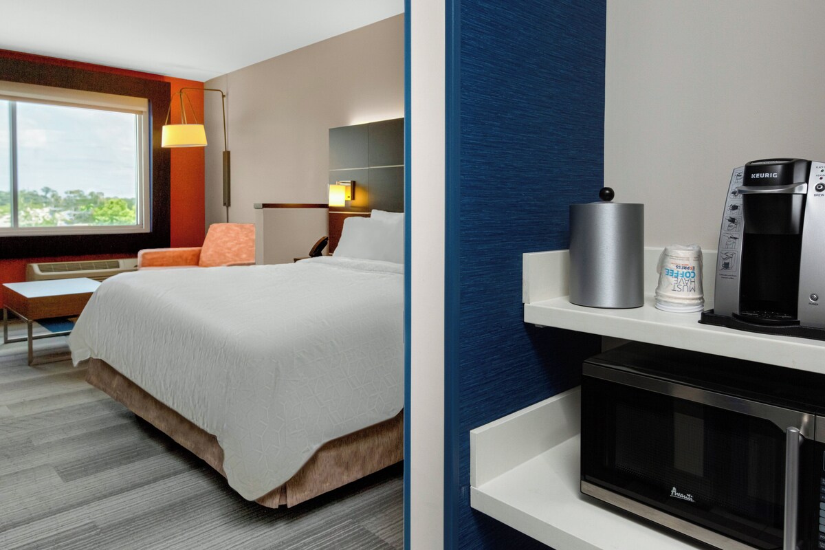 Ein Hotelzimmer mit Bett, Schreibtisch, Stuhl, Lampe, Kaffeemaschine, Mikrowelle und einem Fenster mit Aussicht.