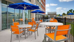 An outdoor patio with round tables and orange chairs, blue umbrellas, and a glass building facade. - Holiday Inn Express & Suites Sanford- Lake Mary by IHG (Sanford)