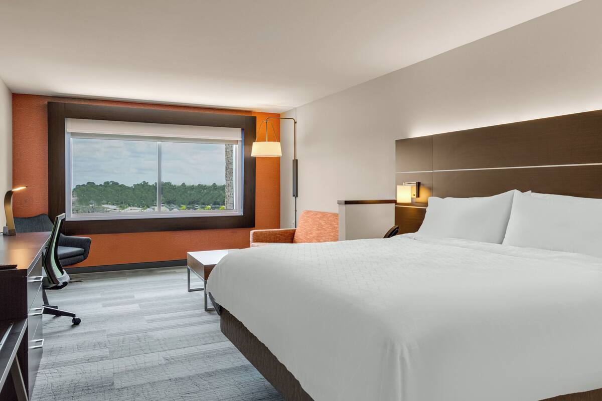 A hotel room with a large bed, a desk with a chair, a nightstand with a lamp, and a view of greenery through a large window.