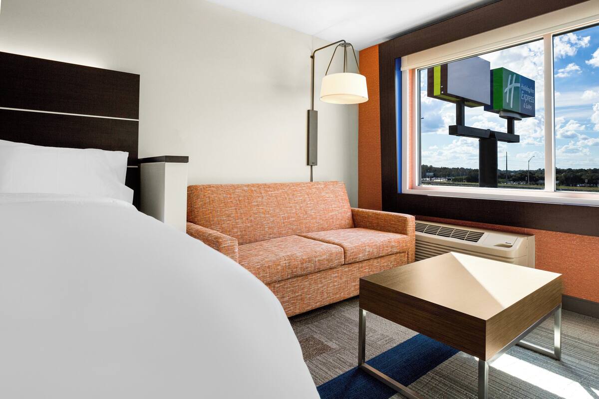 A hotel room with a bed, an orange patterned sofa, a small table, a lamp, and a window with a view of a sign.