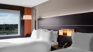 Hotellihuone, jossa on kaksi sänkyä, suuri ikkuna, yöpöytä ja seinään kiinnitetty valaisin.