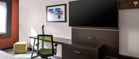 A modern hotel room with a large flat-screen TV, a desk with drawers, a green office chair, and a small ottoman.