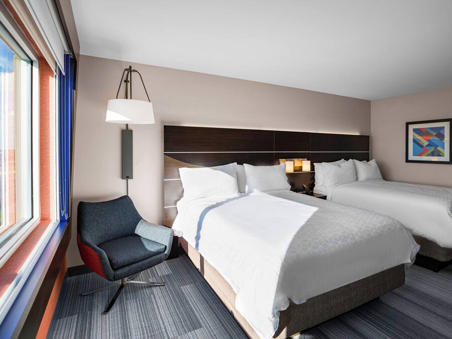 A hotel room with two beds, a chair, a lamp, and a window with blinds.