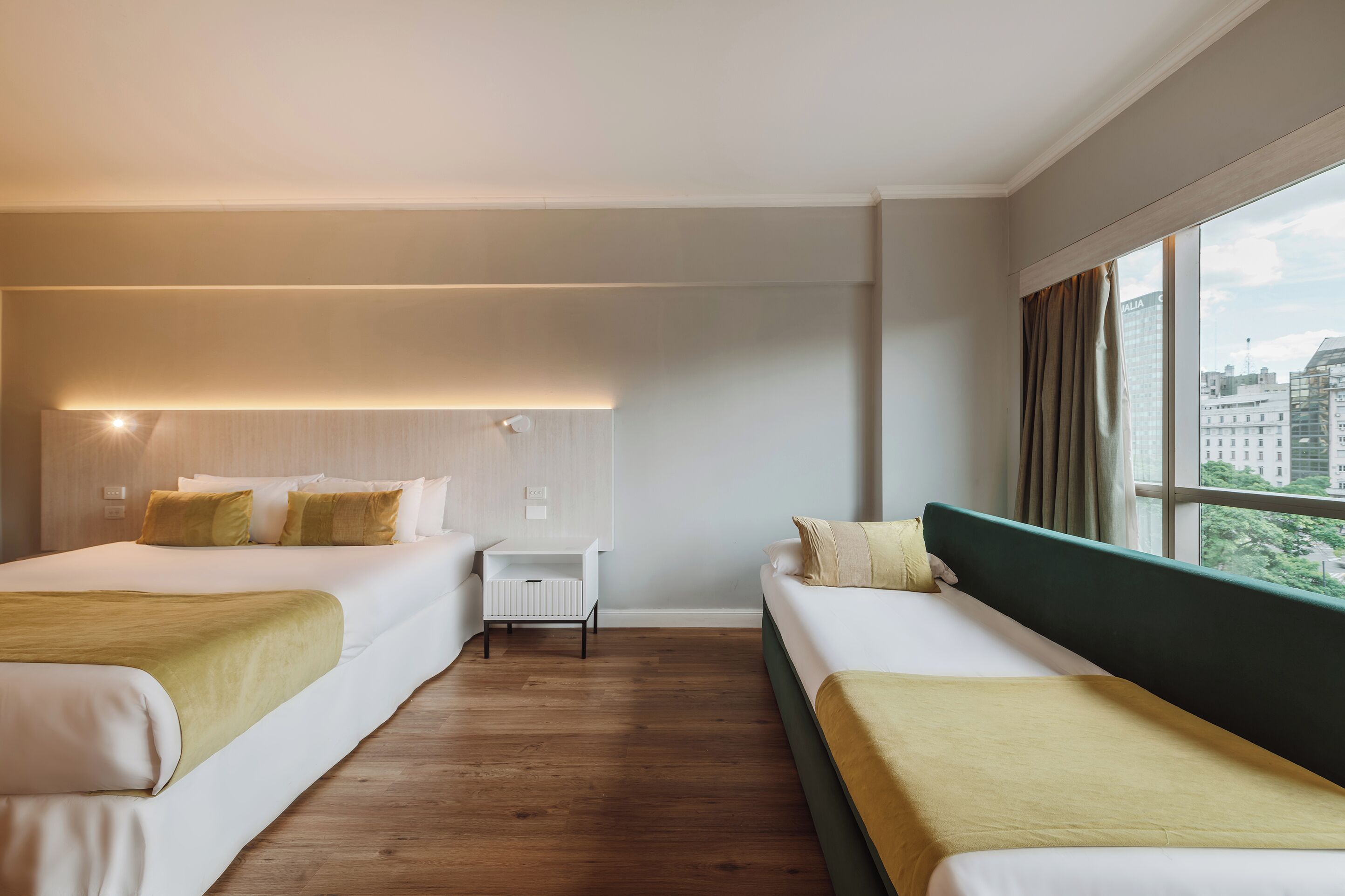 A hotel room with two beds, a small table, and a window with a city view.