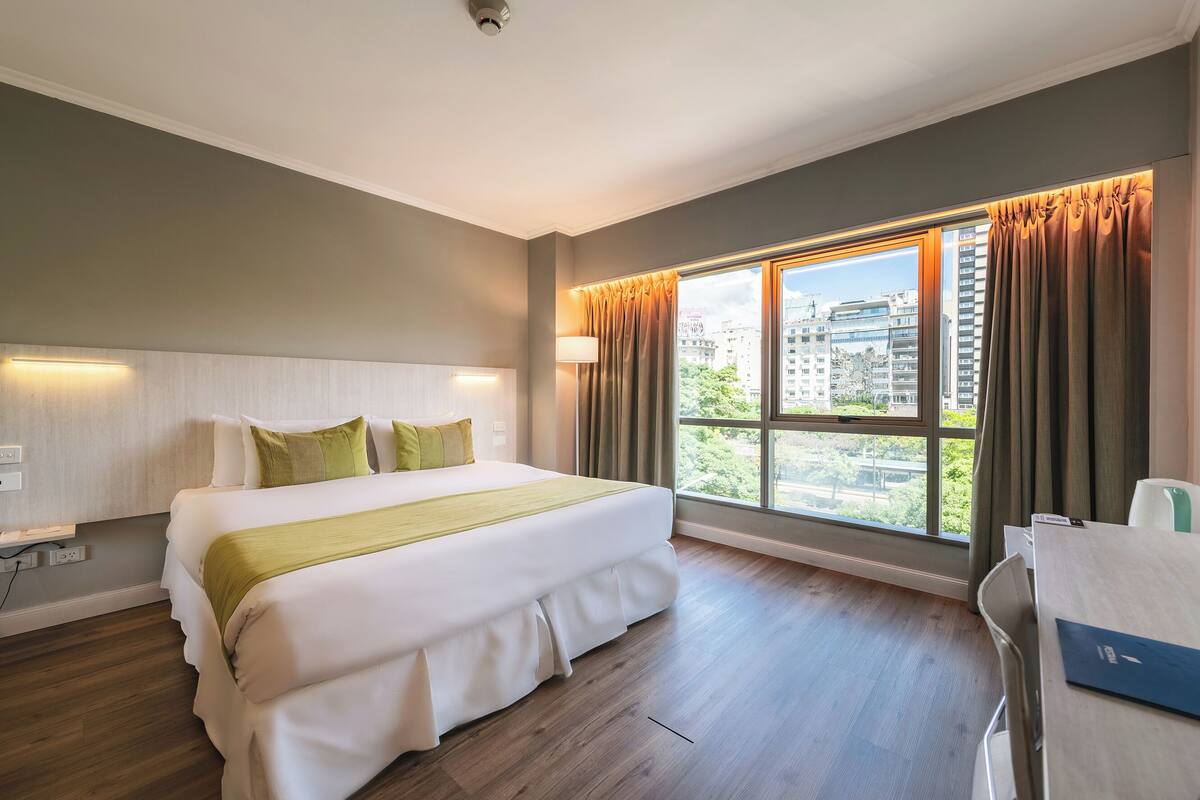 Une chambre d’hôtel avec un grand lit, un bureau et une vue sur la ville grâce à une grande fenêtre.