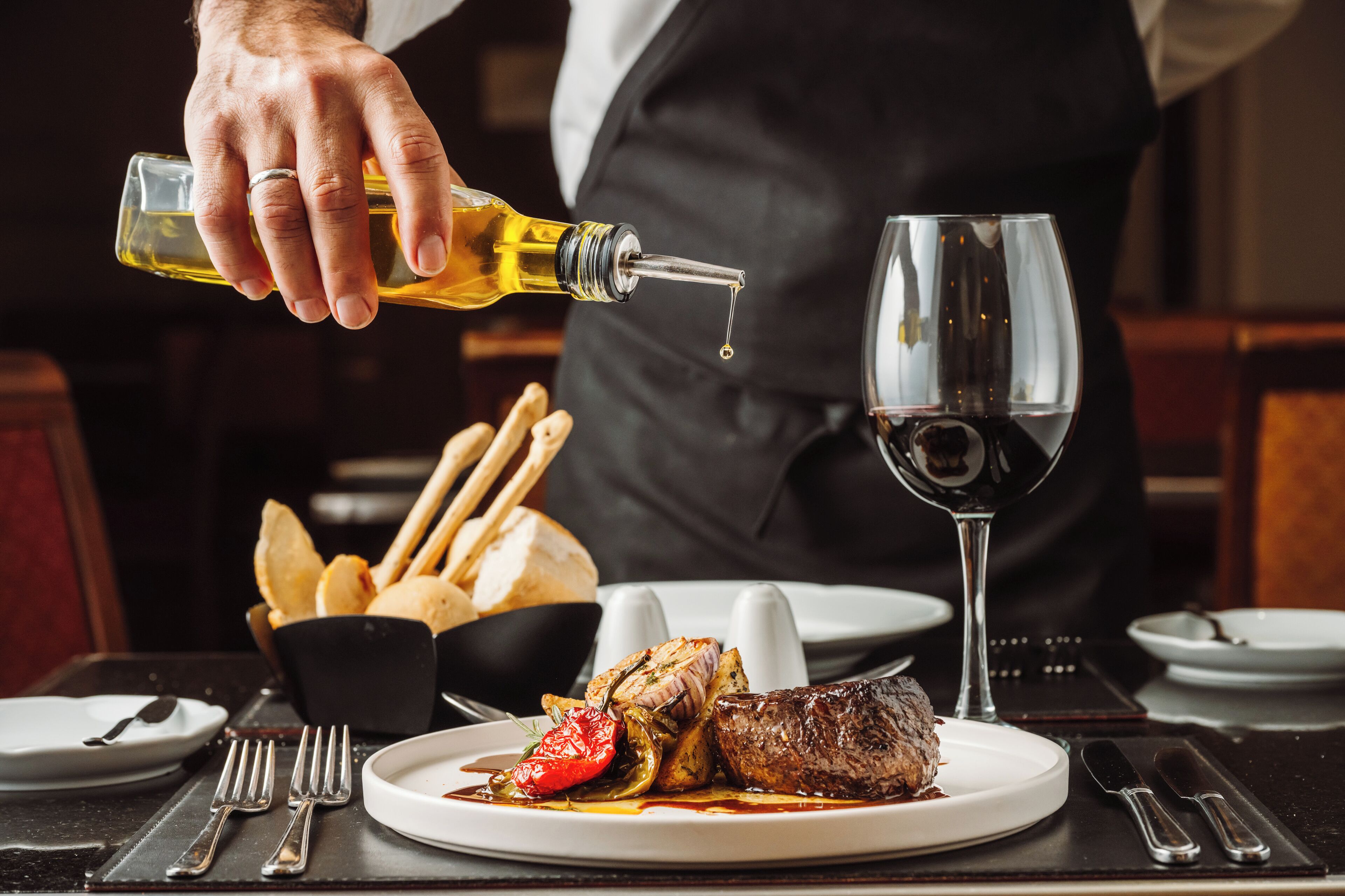 A dining table with a plate of food, a wine glass, and a bottle of olive oil being poured.