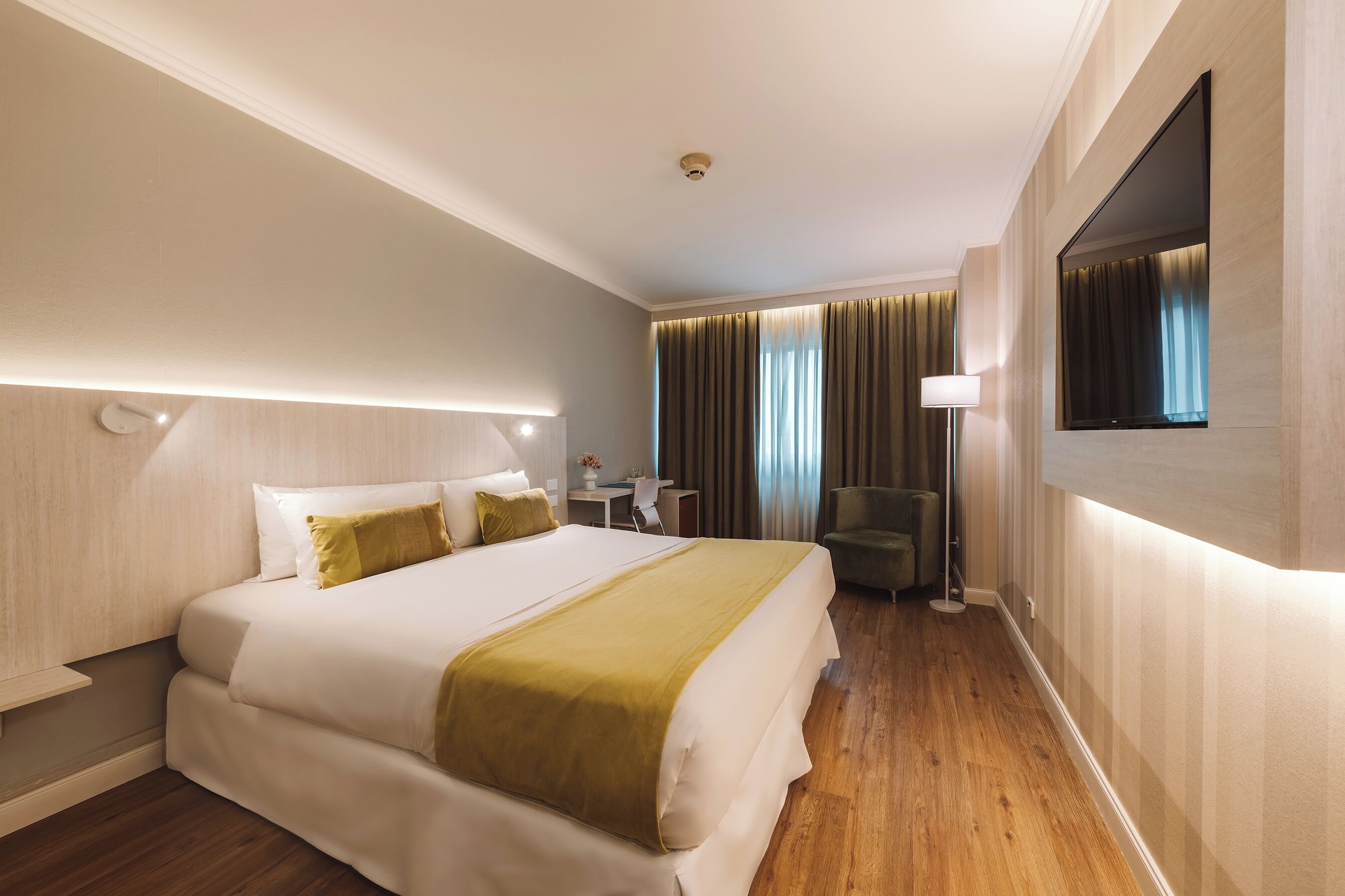 Ein Hotelzimmer mit einem großen Bett, einem Nachttisch, einer Lampe, einem Sessel und einem an der Wand befestigten Fernseher.