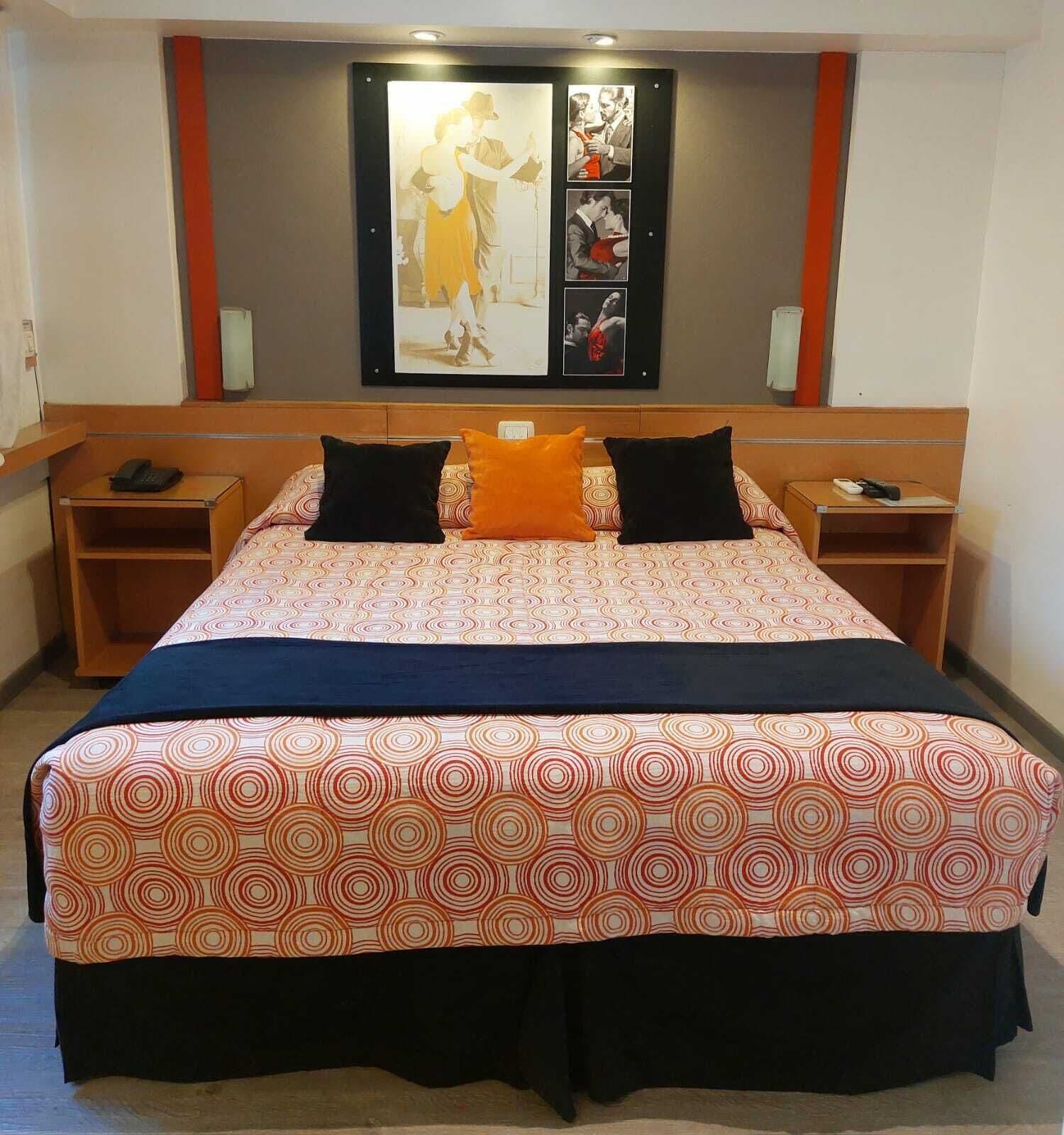 Ein Hotelzimmer mit einem großen Bett, zwei Nachttischen, einem Schminktisch, einem Bettvorhang mit Kunstwerk und einem an der Wand montierten Fernseher.
