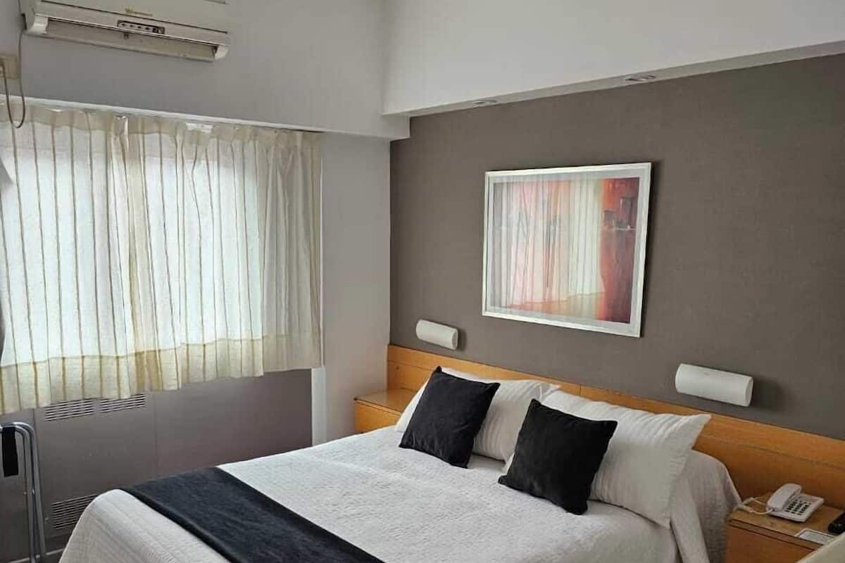 A hotel room with a large bed, a bedside table with a phone, a radiator, and a window with sheer curtains.