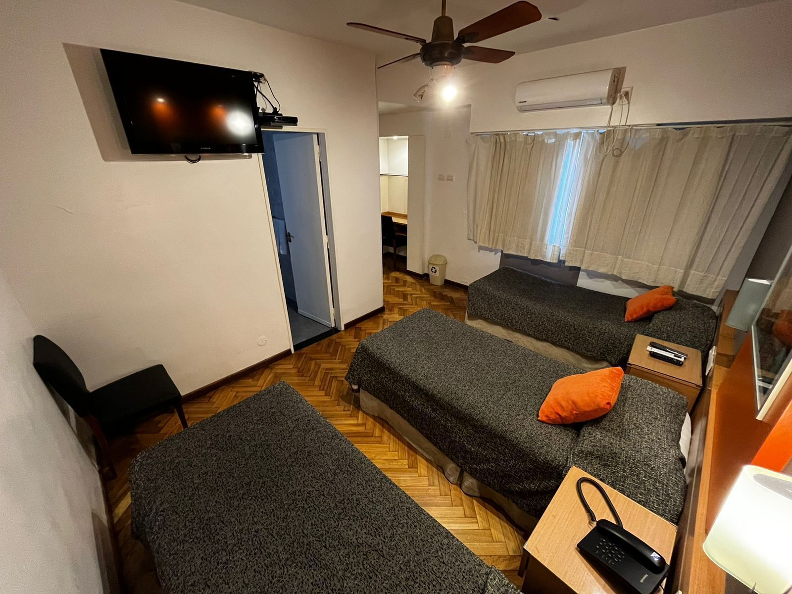 Una habitación con dos camas individuales, un escritorio con silla, un televisor montado en la pared y un ventilador de techo.