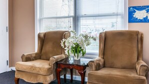 Dos sillones marrones, una mesita de madera y un jarrón con flores blancas frente a una ventana con persianas.