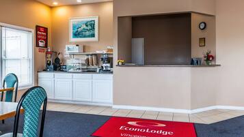 A hotel lobby with a reception desk, a clock, a framed picture, and a dining area with chairs and a table.