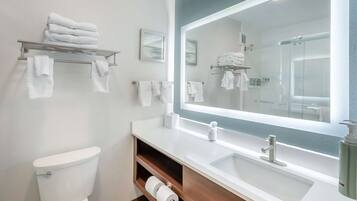Een moderne badkamer met toilet, wastafel en een grote spiegel. Handdoeken liggen op een plankje en er is een douche zichtbaar in de spiegel.