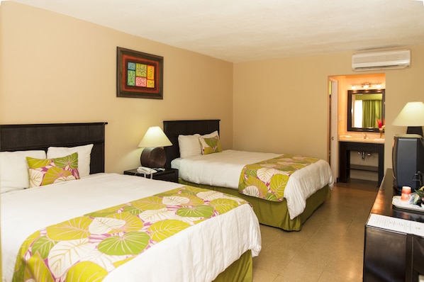A hotel room with two beds, a desk with a lamp, a mirror, and a framed artwork on the wall.