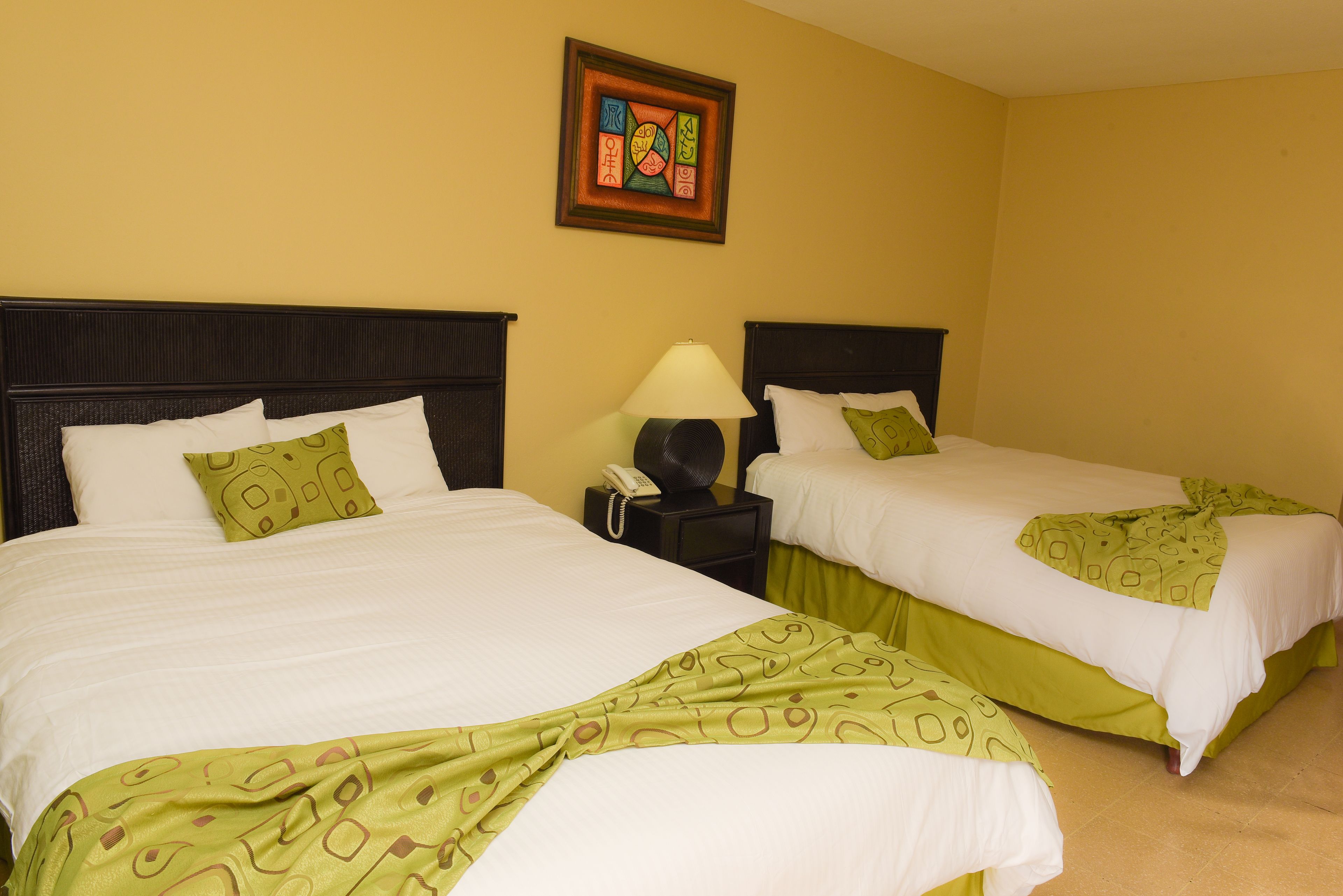 Habitación de hotel con dos camas, cada una con sábanas blancas y colchas estampadas en verde. Entre las camas hay una mesita de noche con una lámpara y un teléfono.