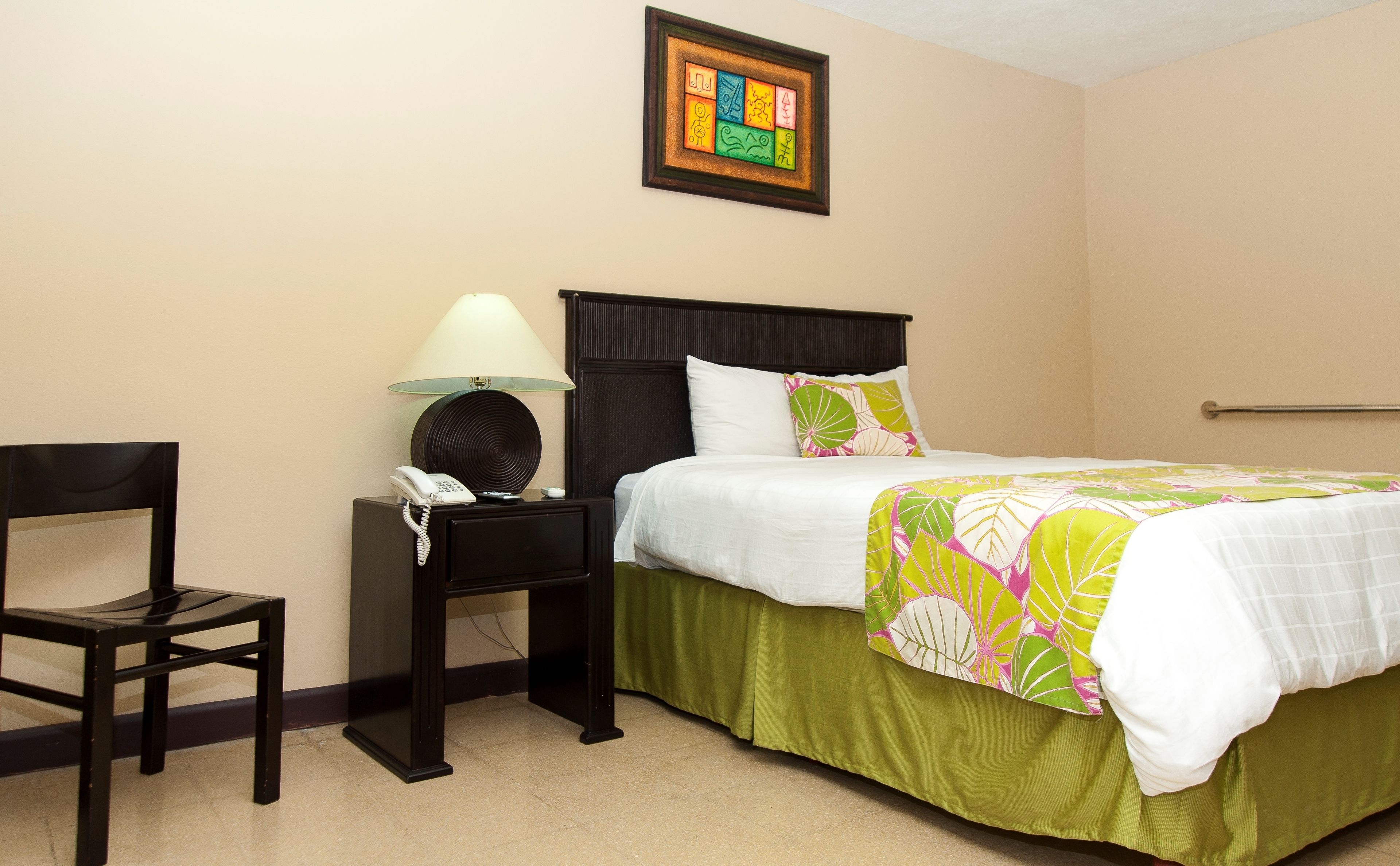 A hotel room with a bed, a chair, a nightstand with a lamp, and a framed artwork on the wall.