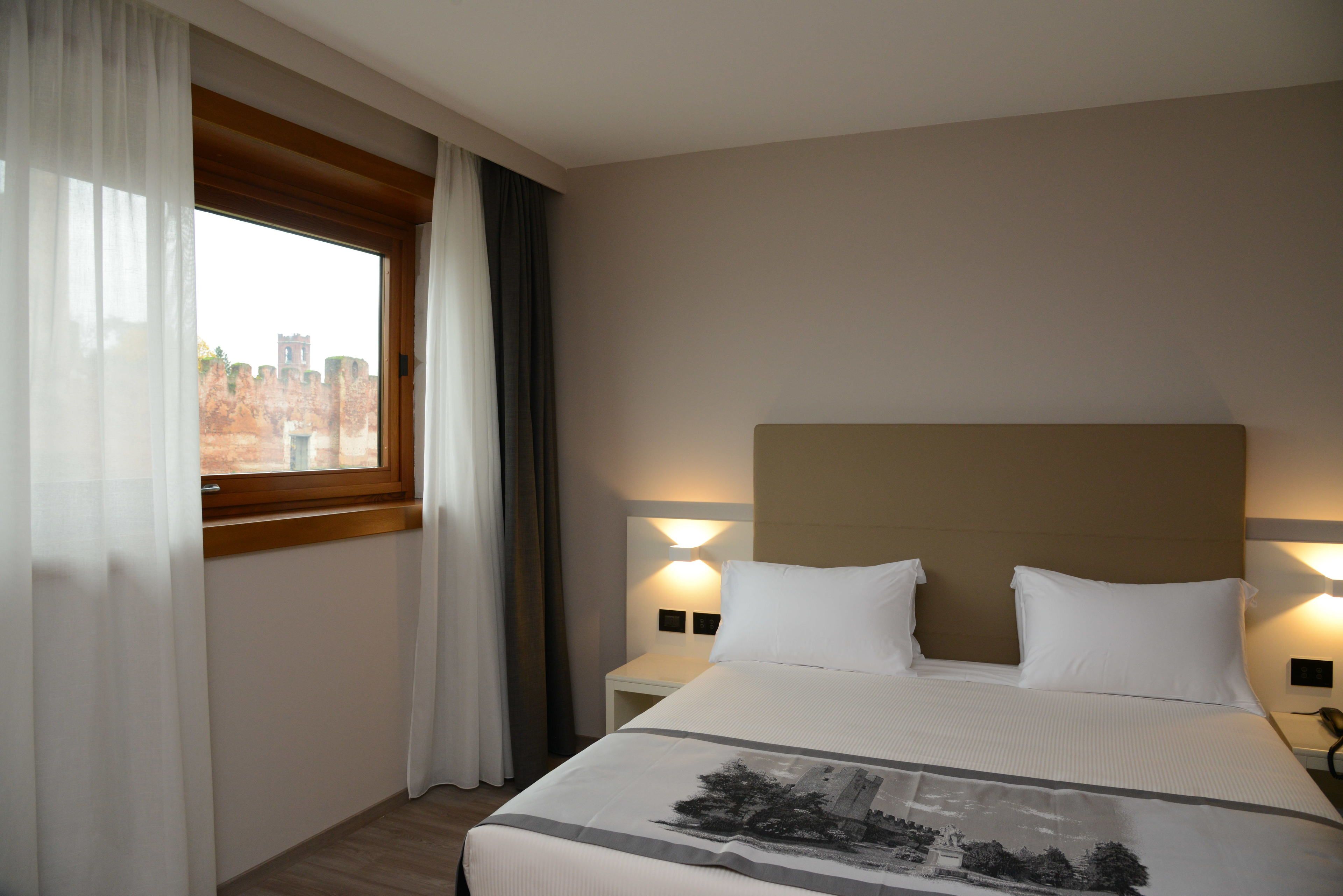 A hotel room with a large bed, a nightstand with a lamp, a window with curtains, and a view of a historic building.