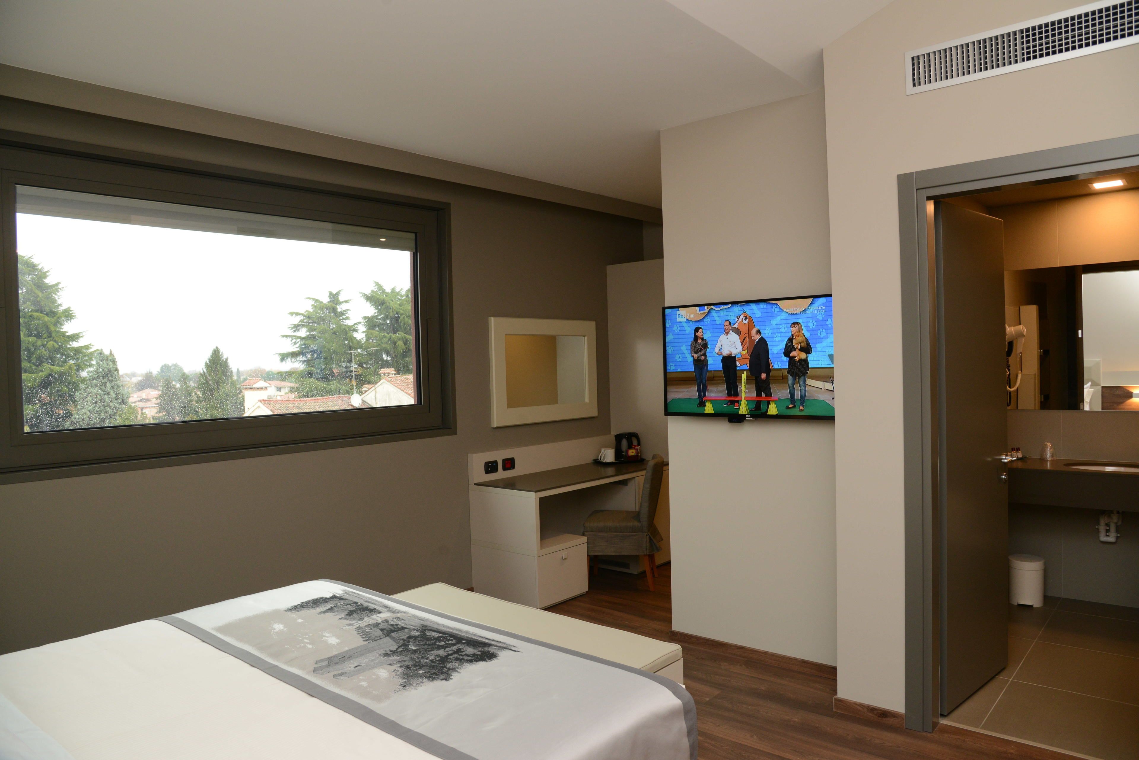 Habitación de hotel con cama, escritorio, televisión y vista a árboles a través de una ventana.