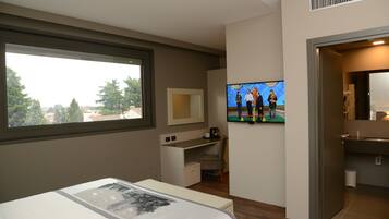 Habitación de hotel con cama, escritorio, televisión y vista a árboles a través de una ventana.