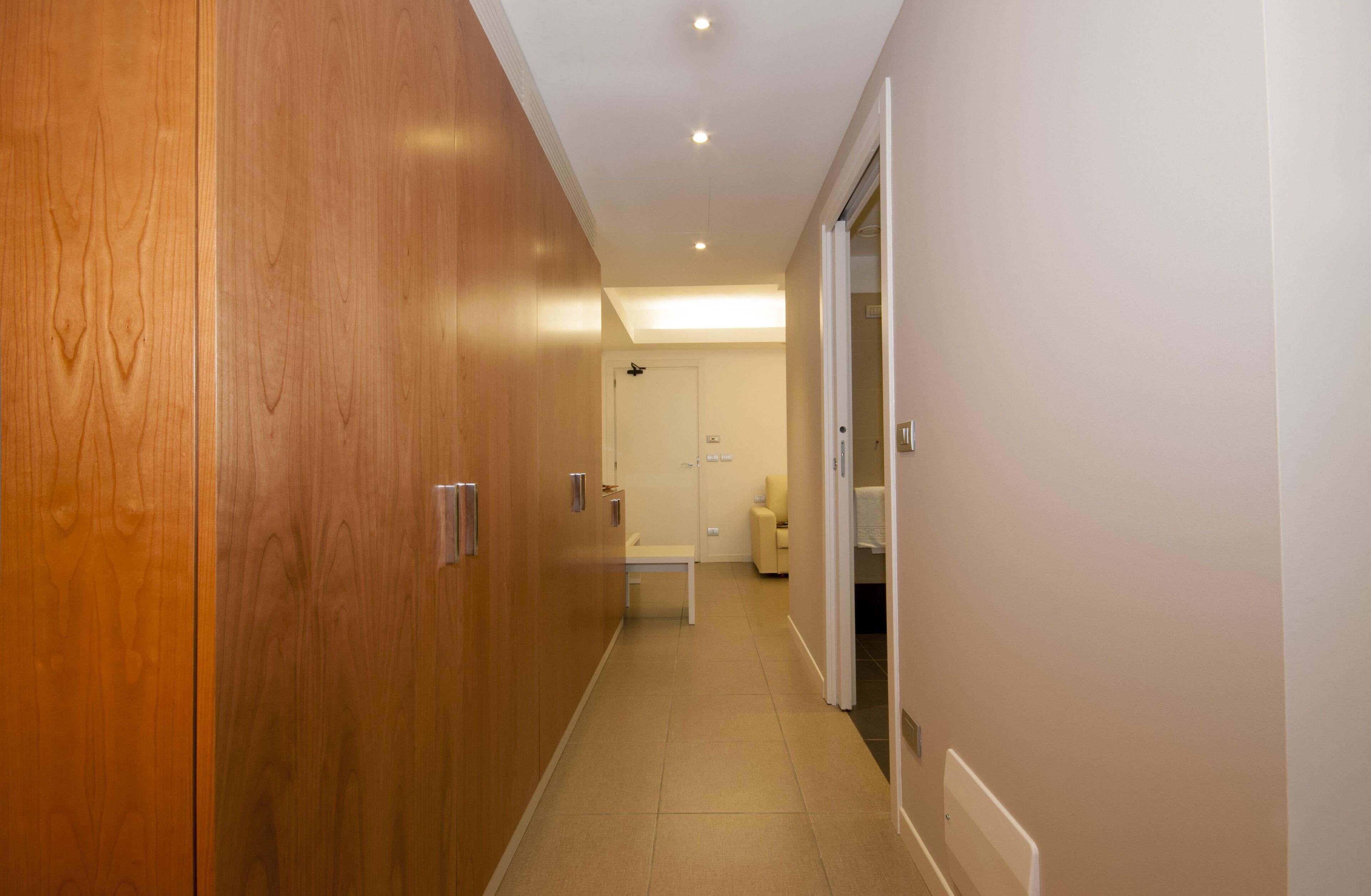 A hallway with wooden doors, a white bench, and a door leading to a room with a bed and a nightstand.