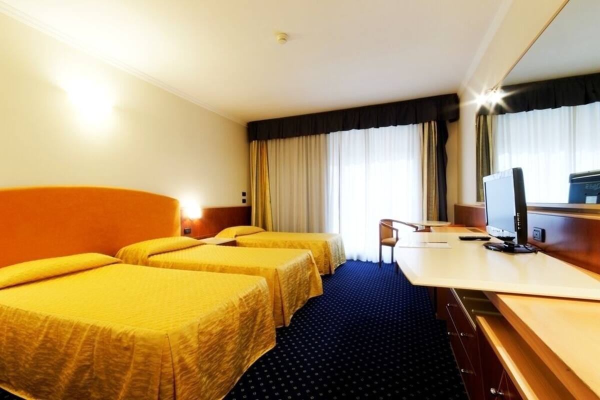 Een hotelkamer met twee eenpersoonsbedden, een bureau met een computer en een groot raam met gordijnen.