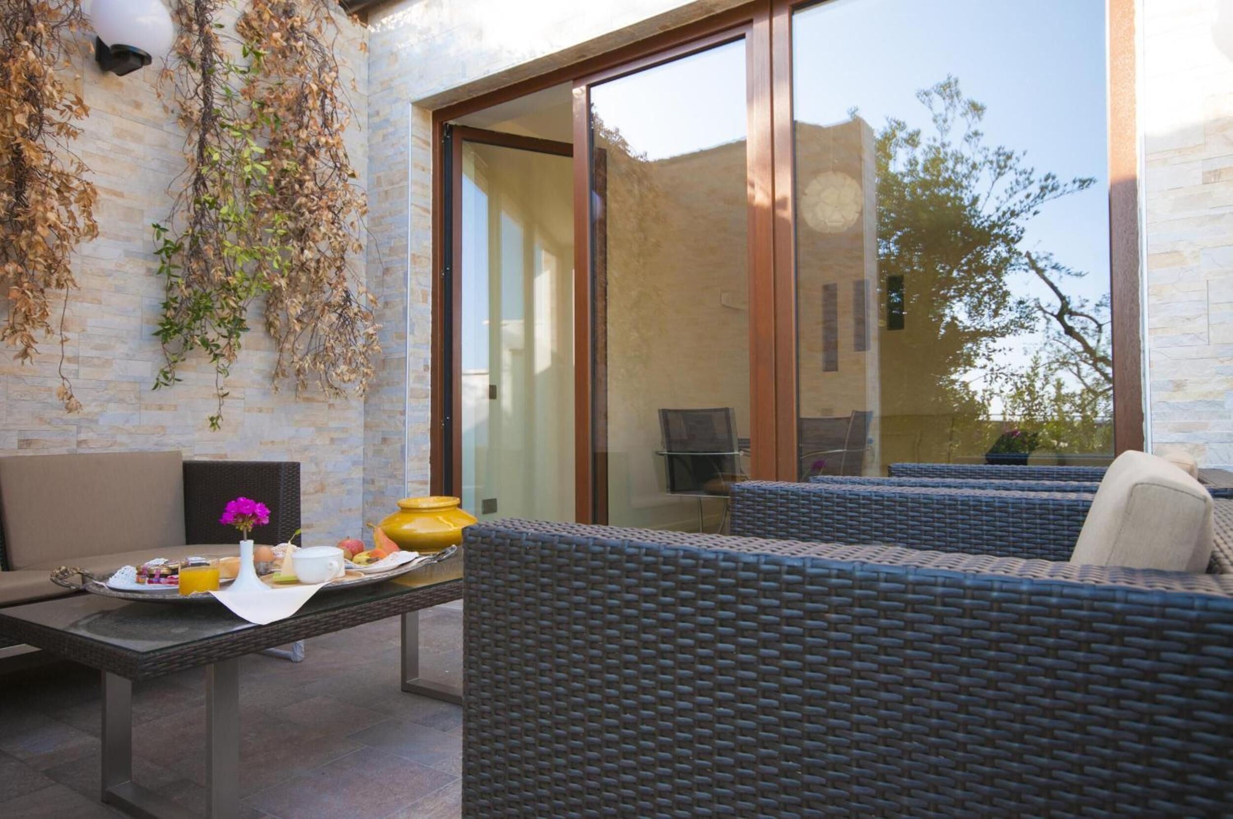 Un espace extérieur aménagé avec des meubles en osier, une table où sont disposés des aliments pour le petit-déjeuner et une porte vitrée donnant sur un patio.