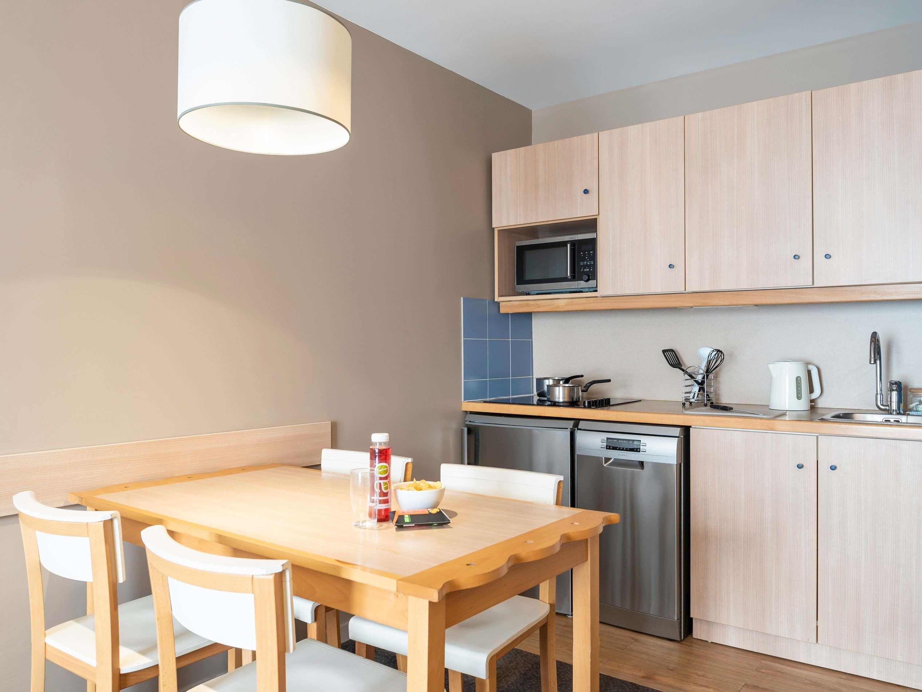 A compact kitchen with a microwave, stove, and dishwasher. There is a wooden dining table with chairs and a hanging light fixture.