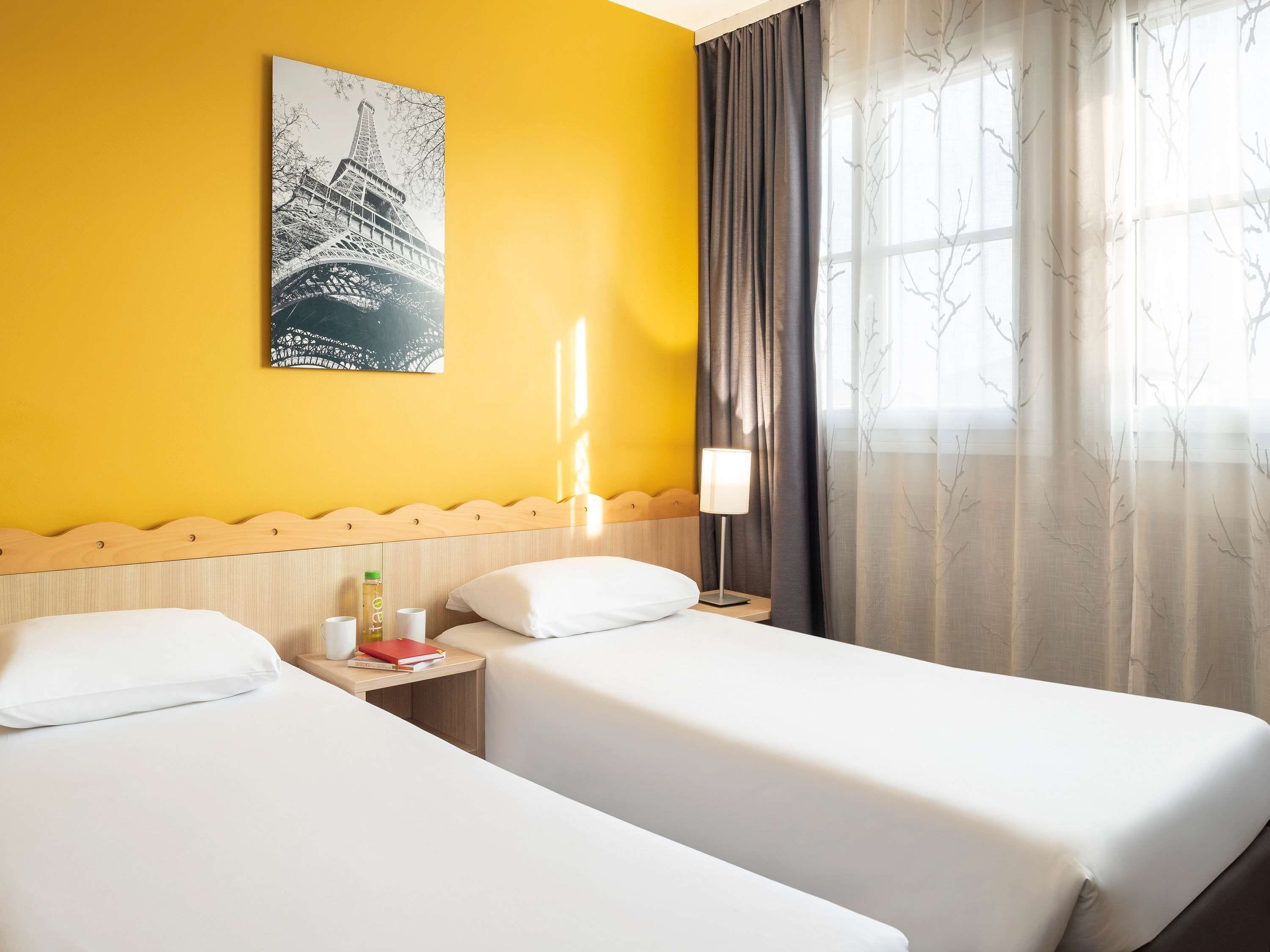 A hotel room with two beds, a wooden headboard, a nightstand with a lamp, a window with sheer curtains, and a cityscape painting on the wall.