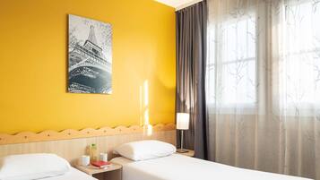 A hotel room with two beds, a wooden headboard, a nightstand with a lamp, a window with sheer curtains, and a cityscape painting on the wall.