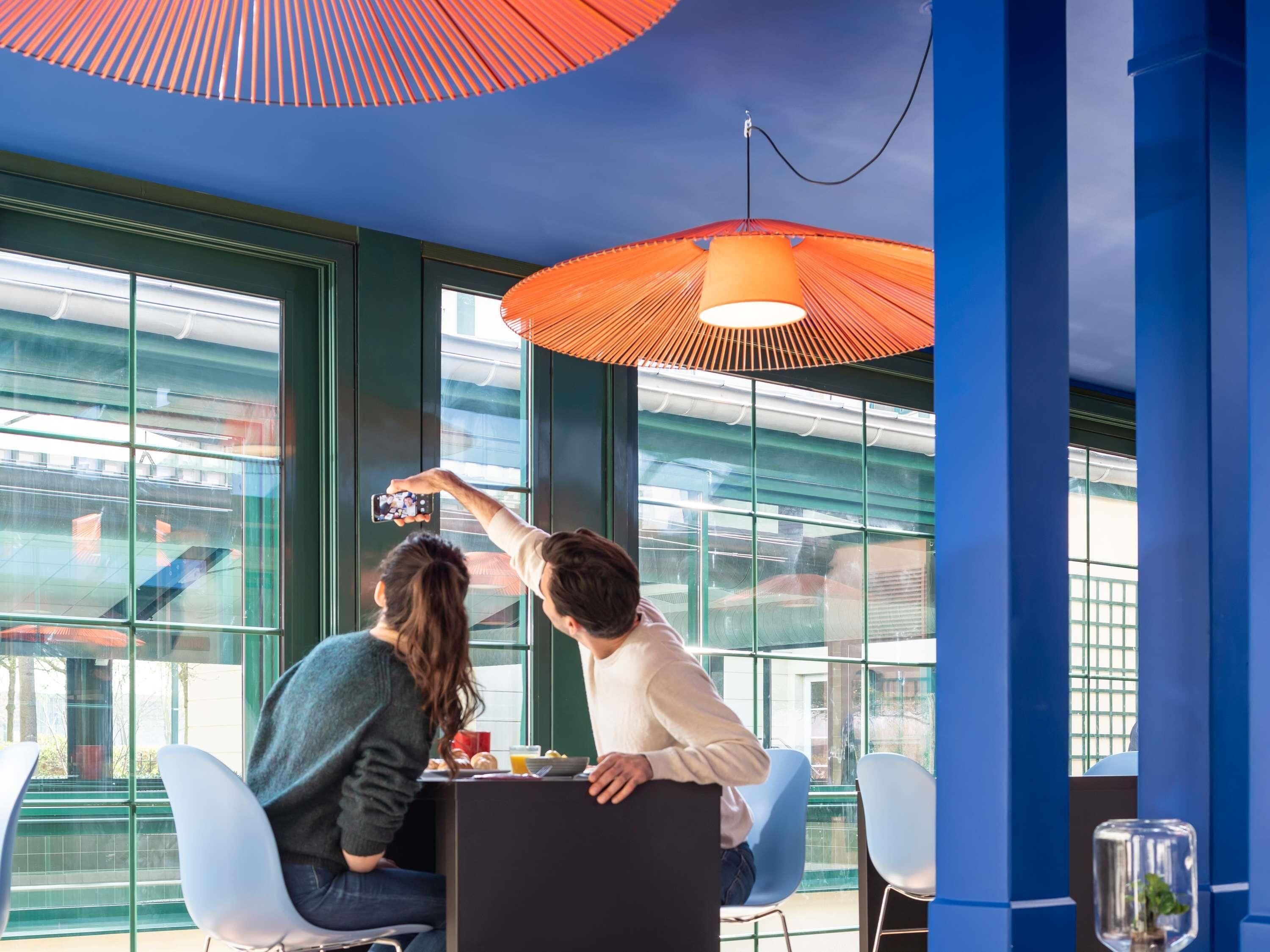 To personer sidder ved et bord med udsigt over byen gennem store vinduer, en rød parasol og moderne indretning.