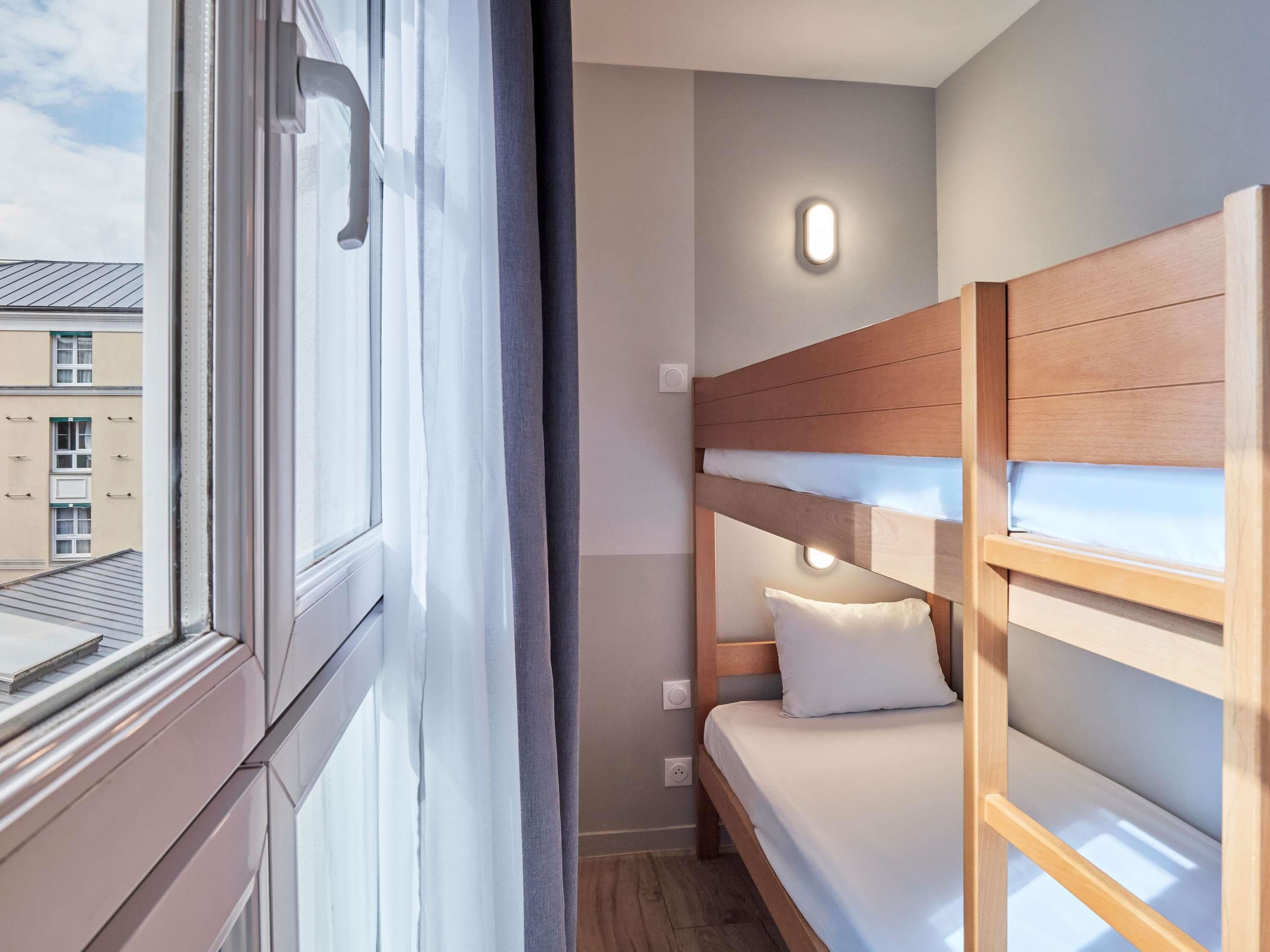 Una habitación de hotel con literas, una ventana con cortinas y una lámpara fijada a la pared.