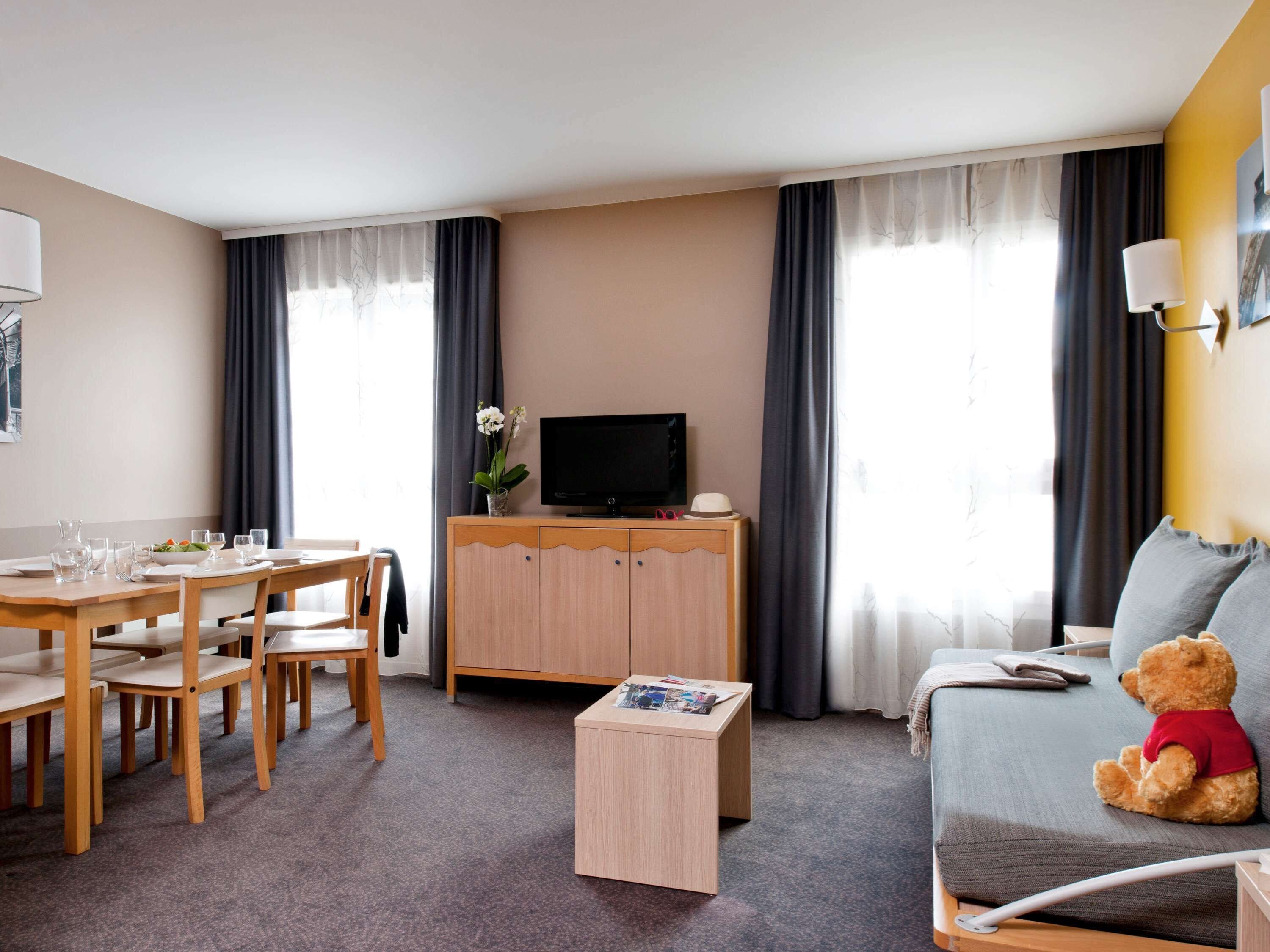 A hotel room with a dining area, a television on a wooden cabinet, a sofa with a teddy bear, and a small table with magazines.