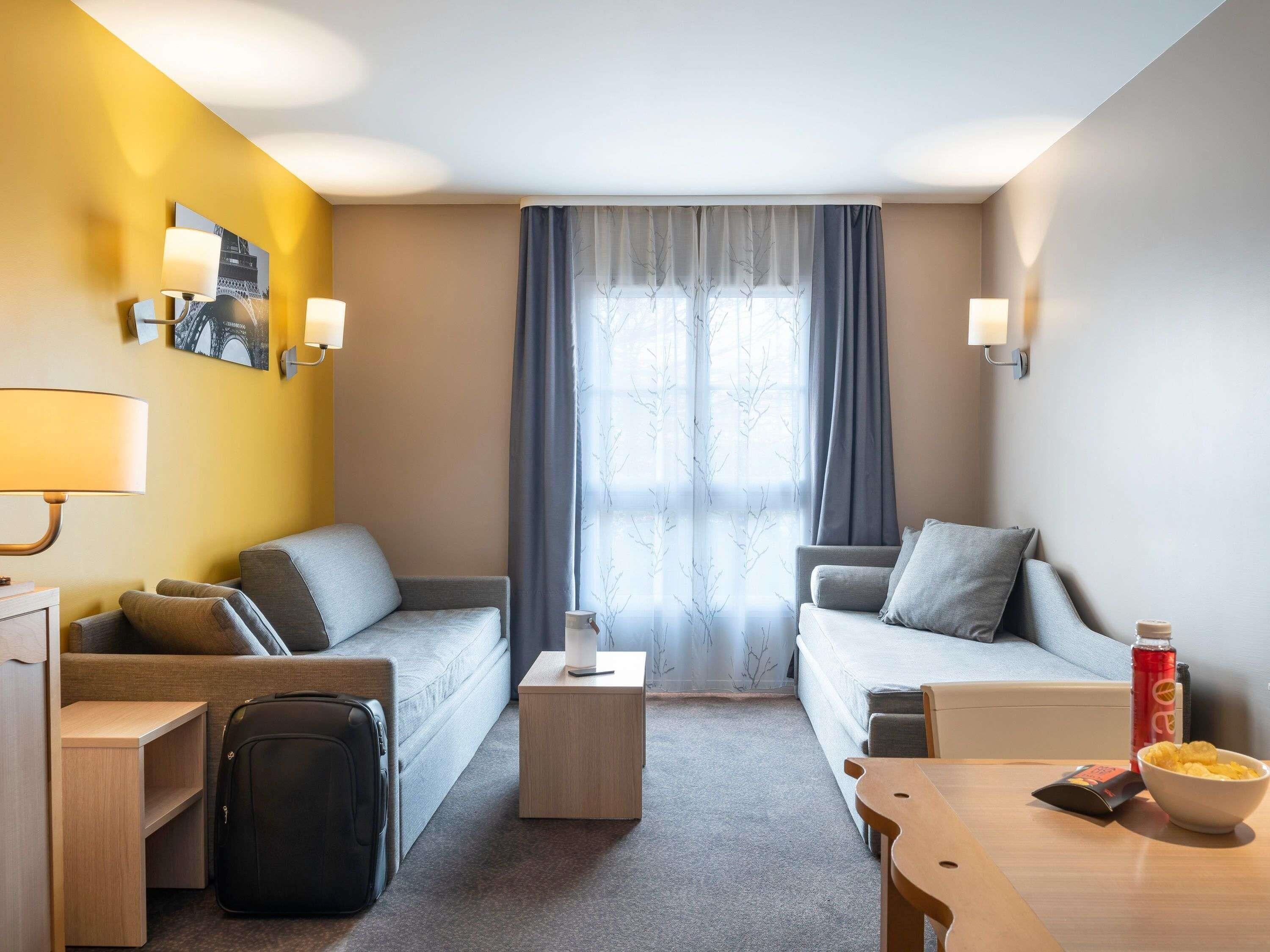 Habitación de hotel con dos sofás grises, una mesita de madera, una lámpara, una maleta y una botella de agua.