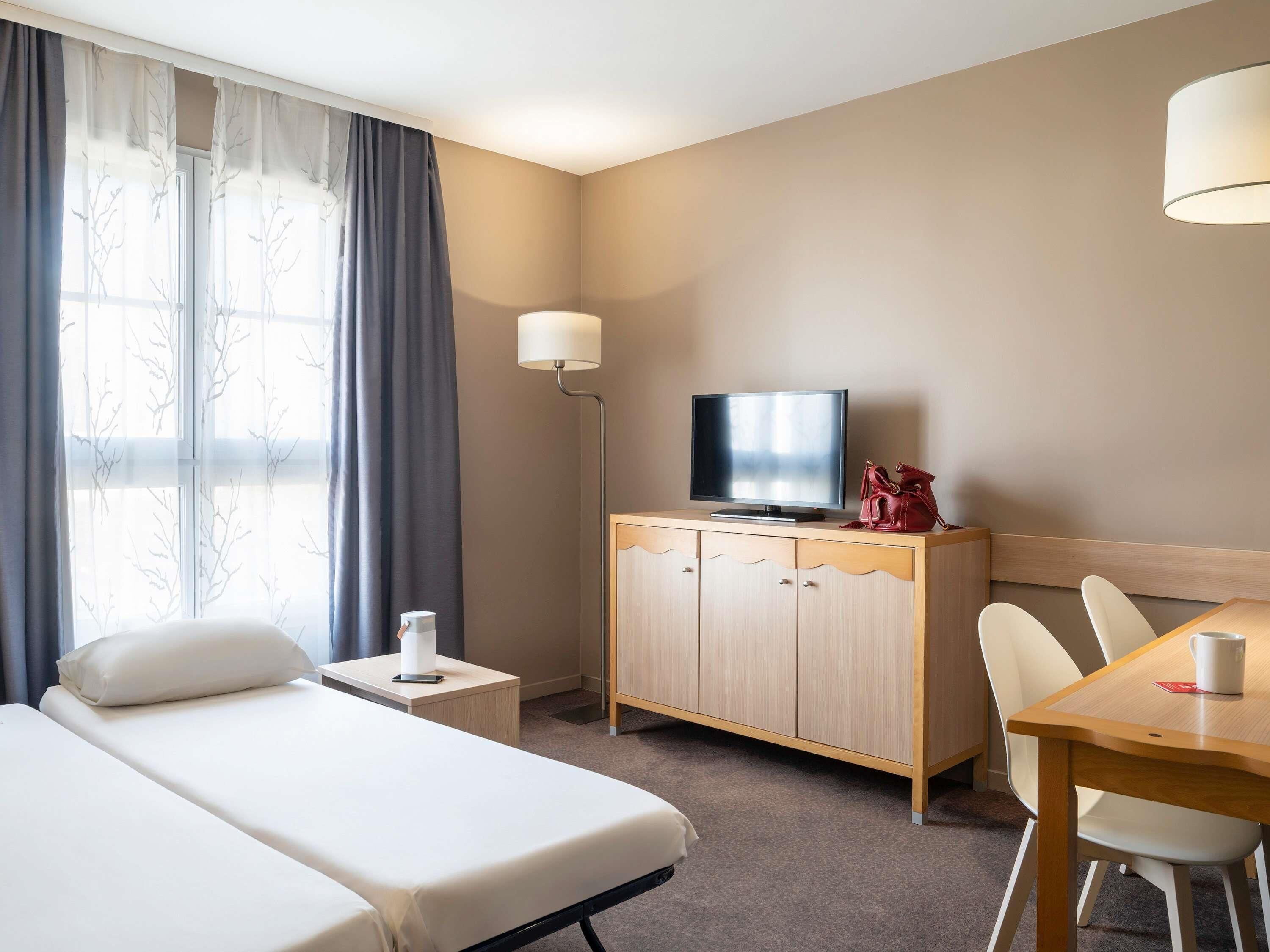 A hotel room with a bed, a TV on a wooden cabinet, a desk with a chair, and a window with curtains.