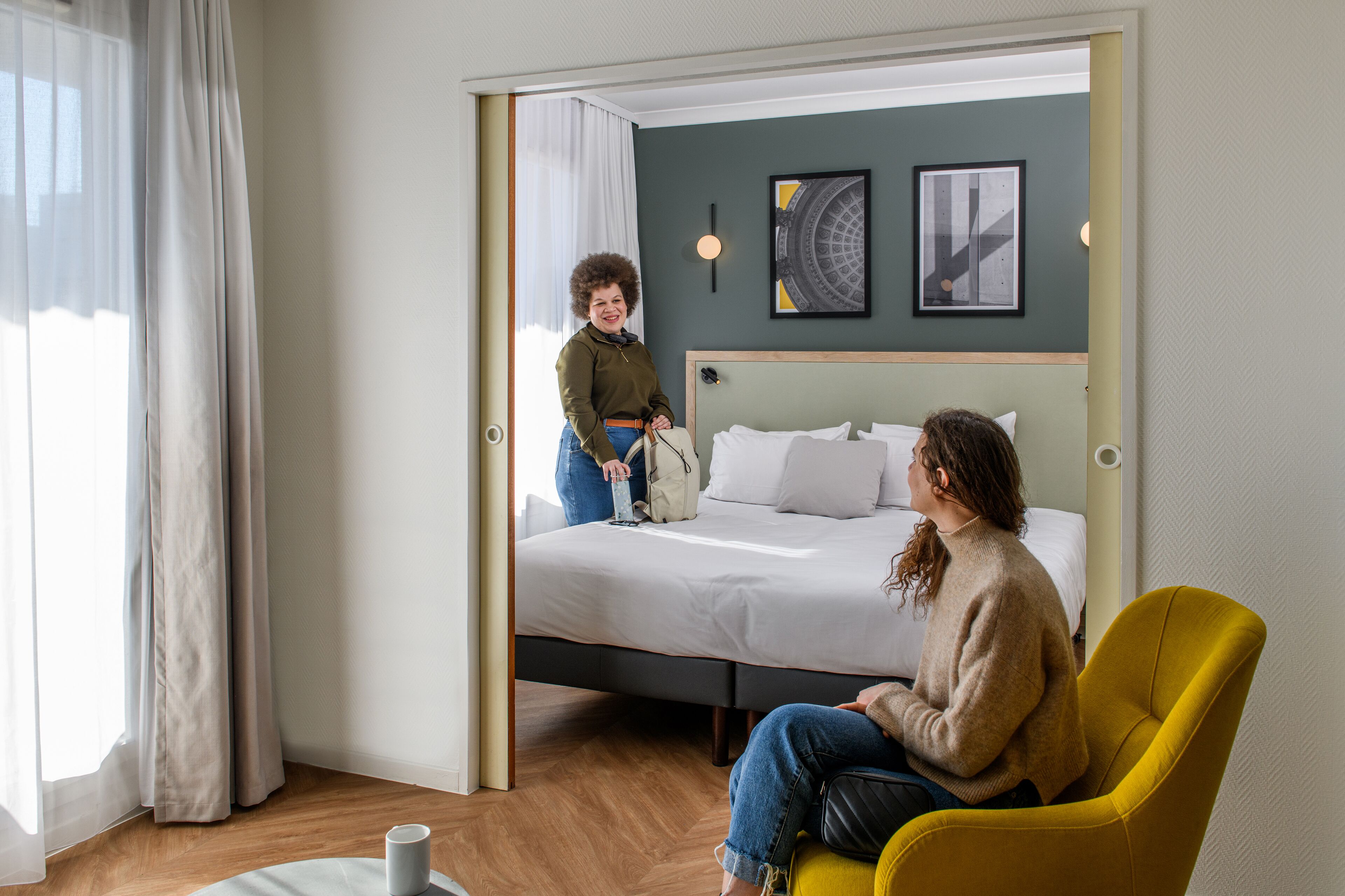 A hotel room with a bed, a yellow armchair, a bedside table with a lamp, and a window with curtains.