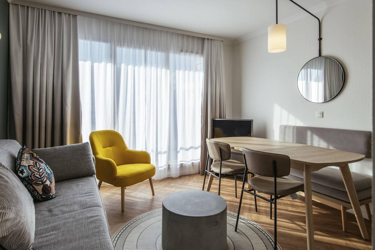 A modern living room with a sofa, yellow armchair, dining table, and a round grey ottoman.