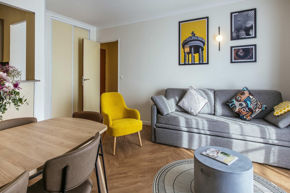 Una sala de estar moderna con una mesa de comedor, un sillón amarillo brillante, un sofá gris con cojines estampados y una mesa redonda de centro.