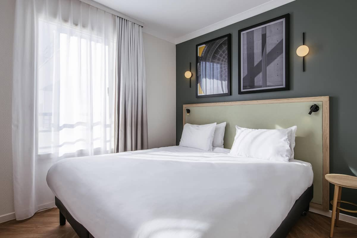 Habitación de hotel con una cama grande, cabecero de madera, un taburete pequeño de madera y dos cuadros en la pared.