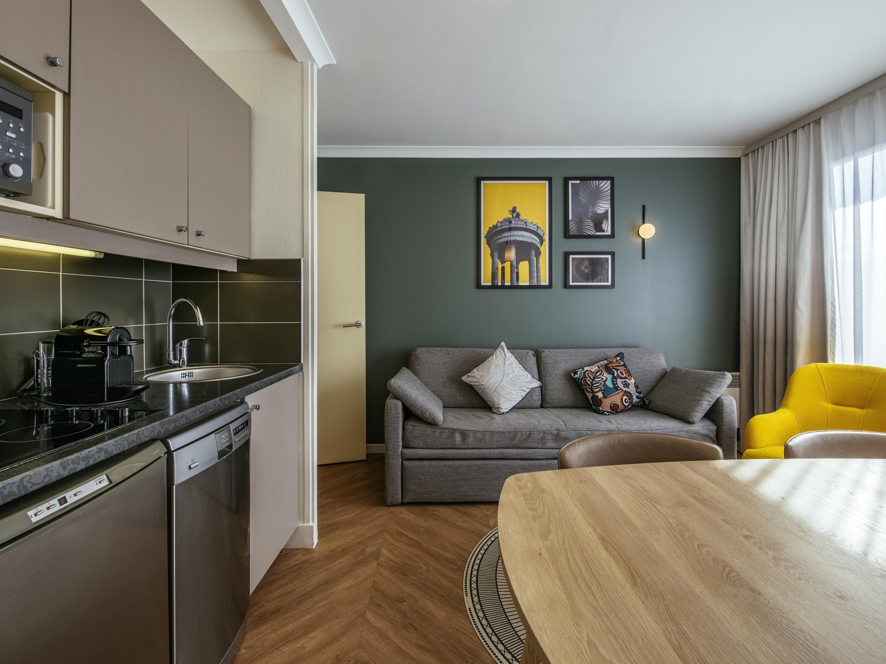 A modern kitchen with a dishwasher, a coffee machine, and a sink. There is a grey sofa with patterned cushions, a wooden dining table, and a yellow armchair. The room has a green accent wall with framed artwork.