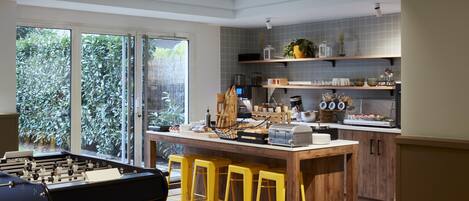 A modern kitchen with a foosball table, bar stools, and a view of greenery through large windows.