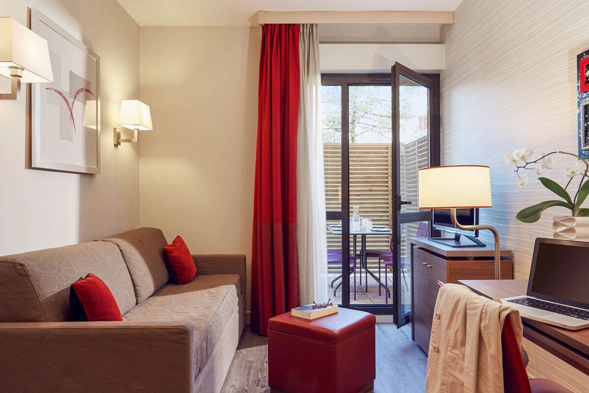 A hotel room with a sofa, a desk with a laptop, a small red ottoman, and a view of the outdoor area through a glass door.